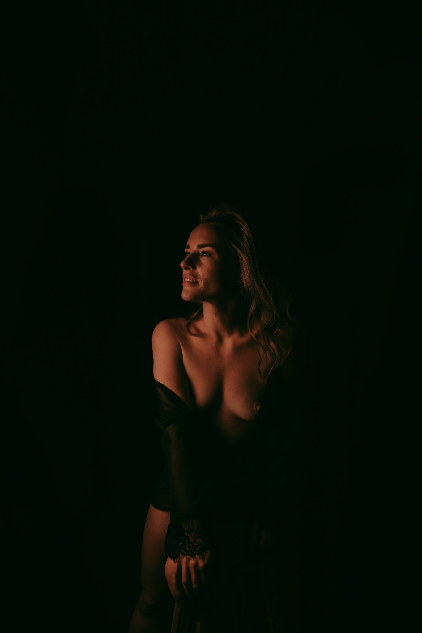 Donker portretfoto waarbij een vrouw met blonde haren op een krukje zit en ze heeft een zwarte kimono aan, de kimono valt over haar schouder