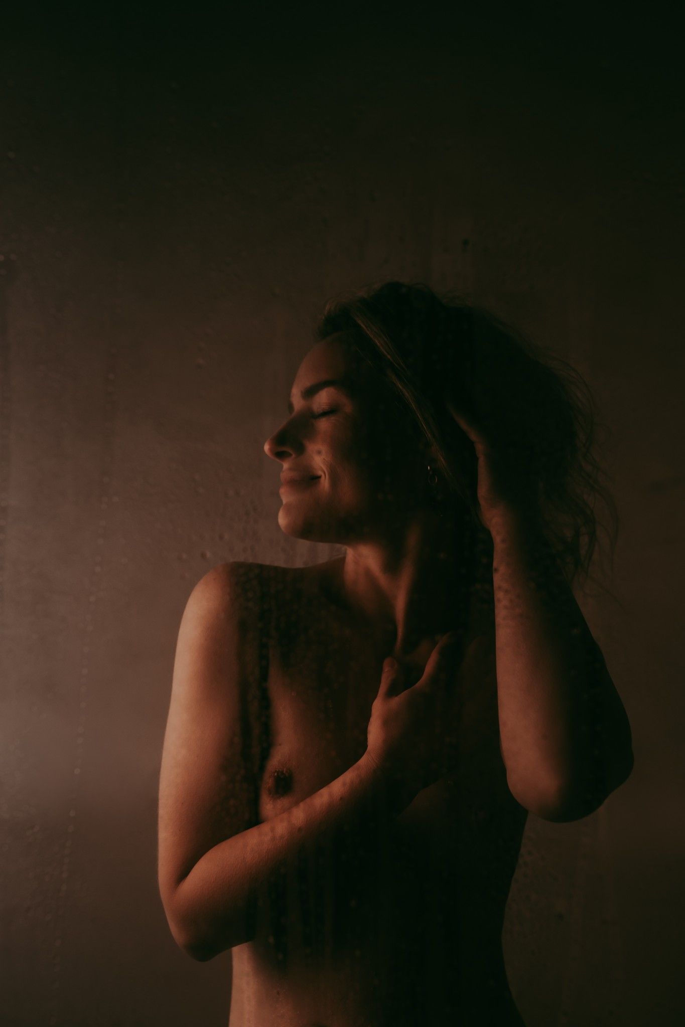Portret foto van een vrouw die haar haren vast houdt en naar de zijkant kijkt met een kleine lach. Staand voor een raam met druppels