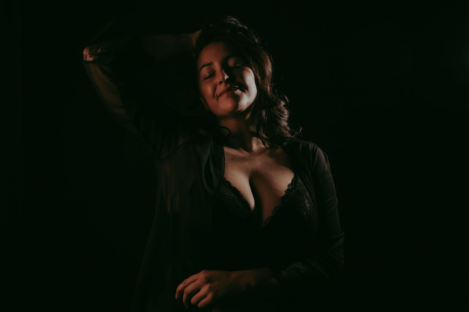 Foto van een vrouw met lingerie en een kimono in het zwart met een donkere zwarte achtergrond geposeerd met 1 arm houdt ze haar haren vast.