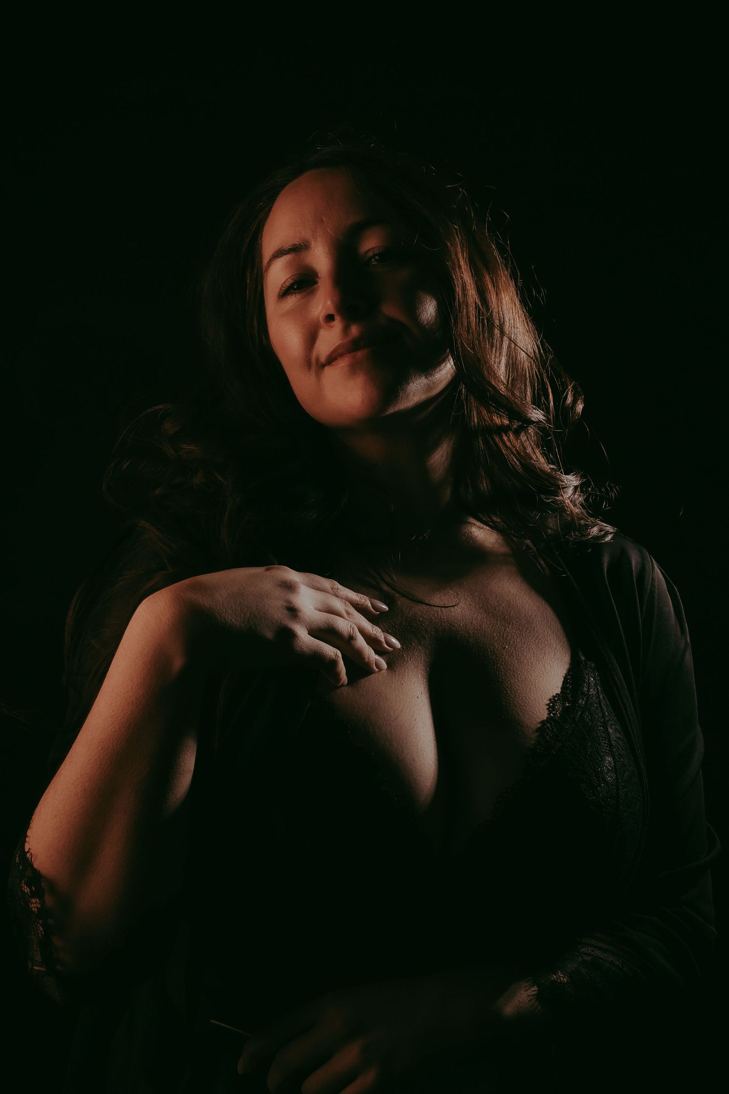 A woman in dark lingerie poses dramatically against a black background in a moody boudoir portrait.