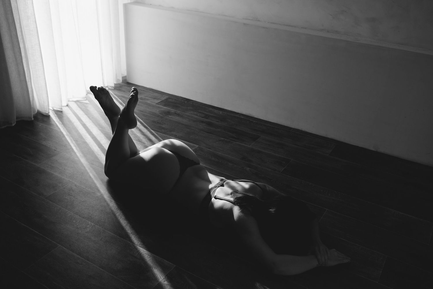 Black and white photo of a person lying on hardwood floor near a window with soft natural light.