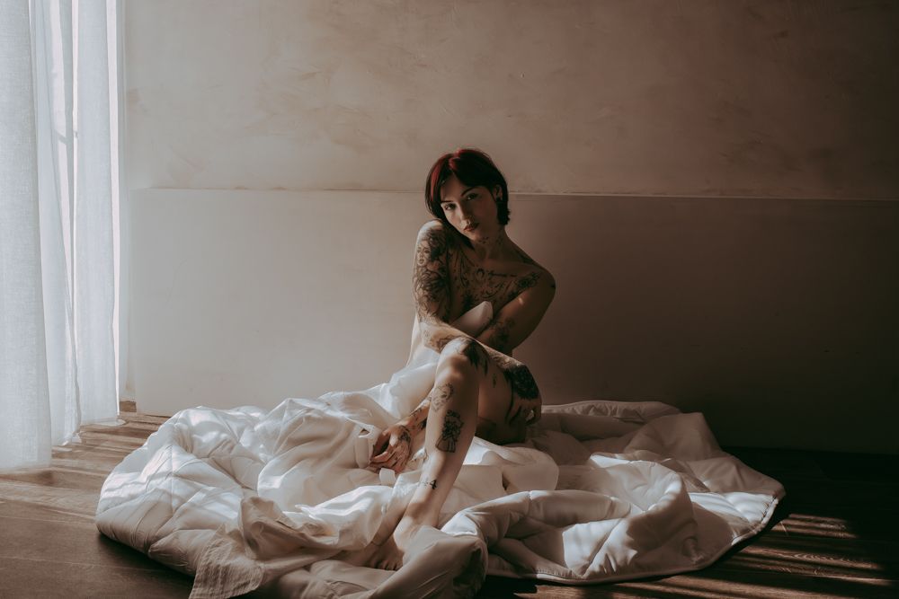 A moody boudoir photograph shows a figure sitting on rumpled white bedding in a minimalist room with natural light.