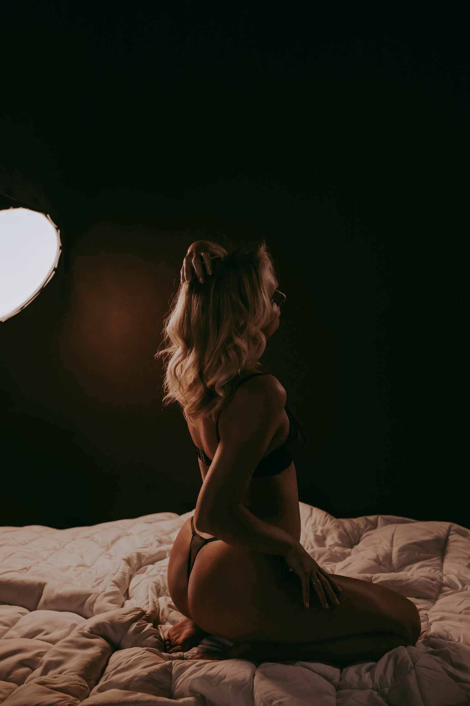 A blonde woman in black lingerie kneels on a white bed, illuminated by a studio light against a dark background.