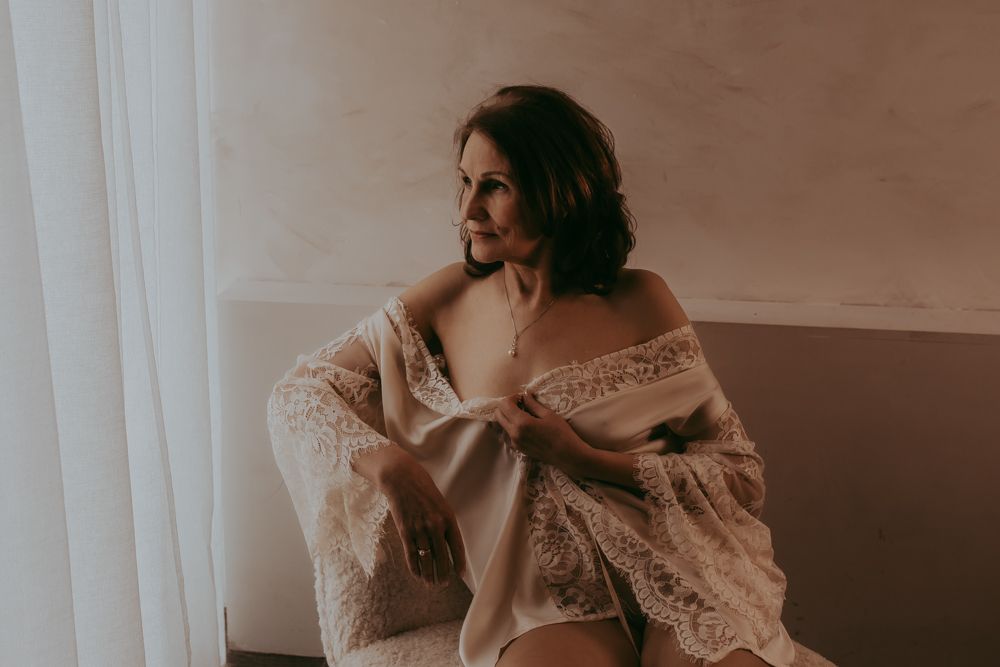 Woman in white lace dress lounges on furniture in romantic boudoir photo session.
