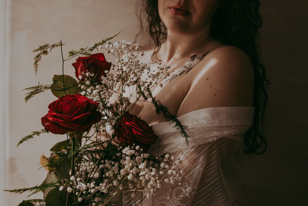 A romantic boudoir photo showing a sheer white lace covered figure holding vibrant red roses and baby's breath.