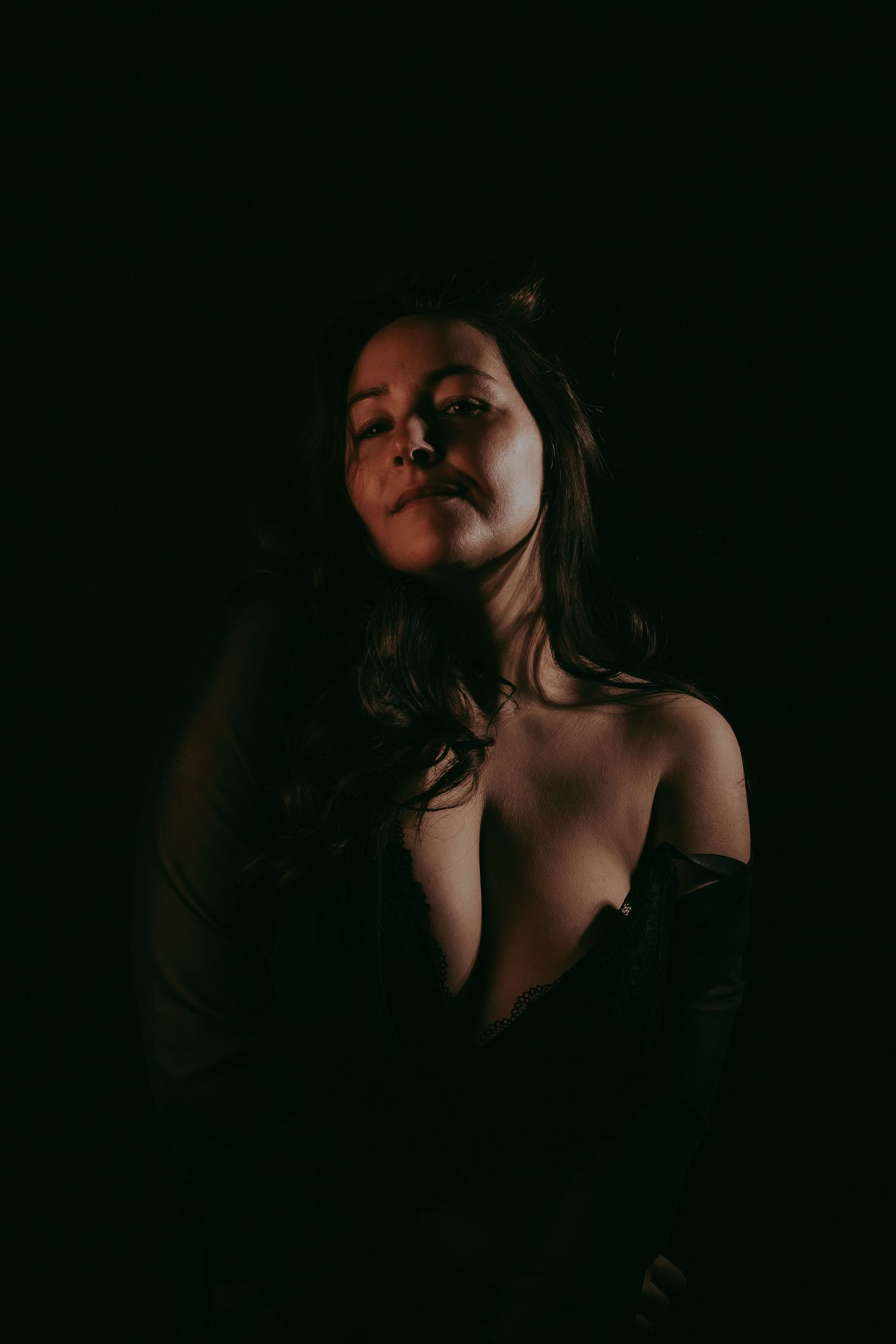 A woman with long dark hair poses dramatically in low-key lighting against a dark background, wearing an off-shoulder dark outfit.