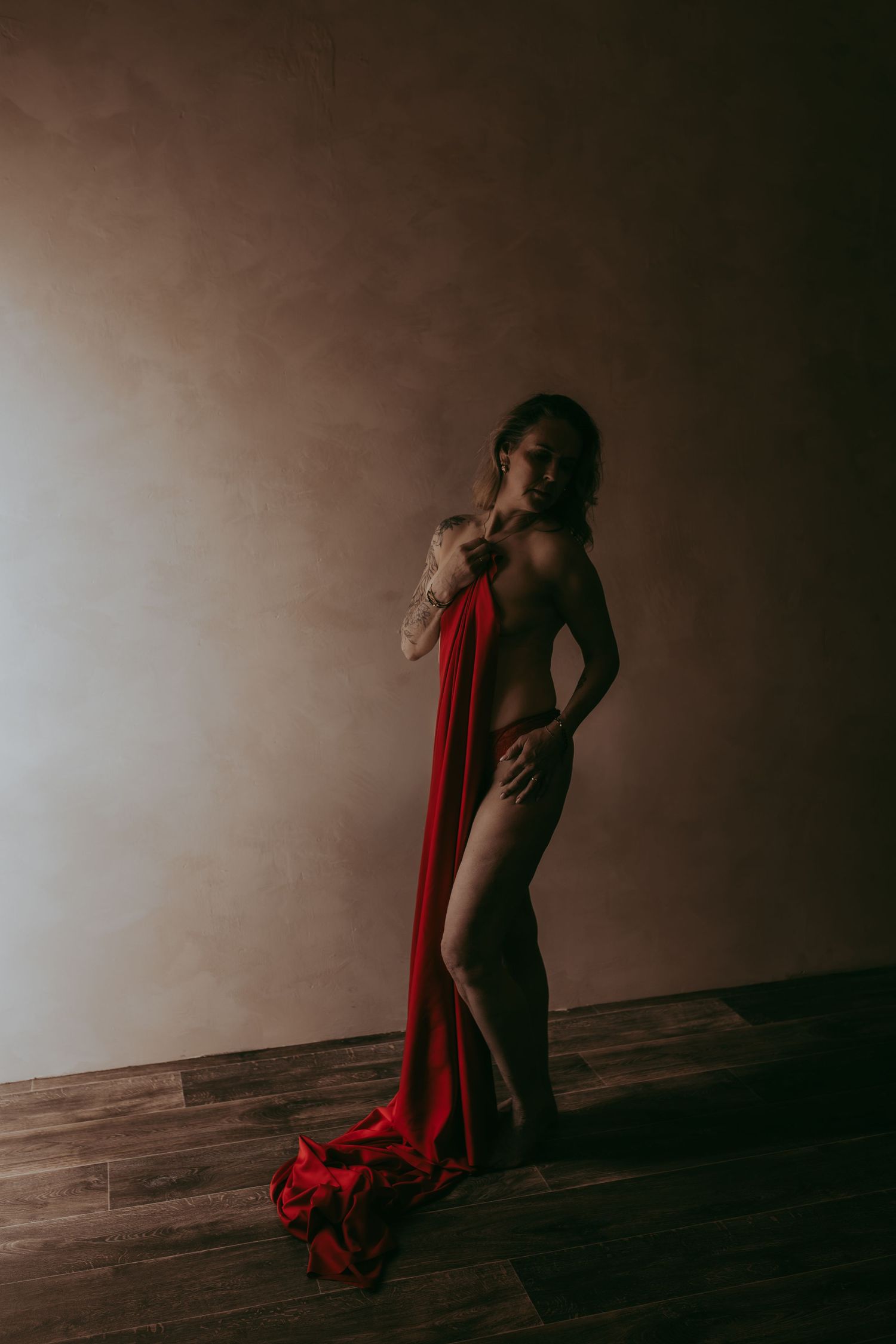 A woman poses dramatically with flowing red fabric against a moody studio backdrop on hardwood floors.