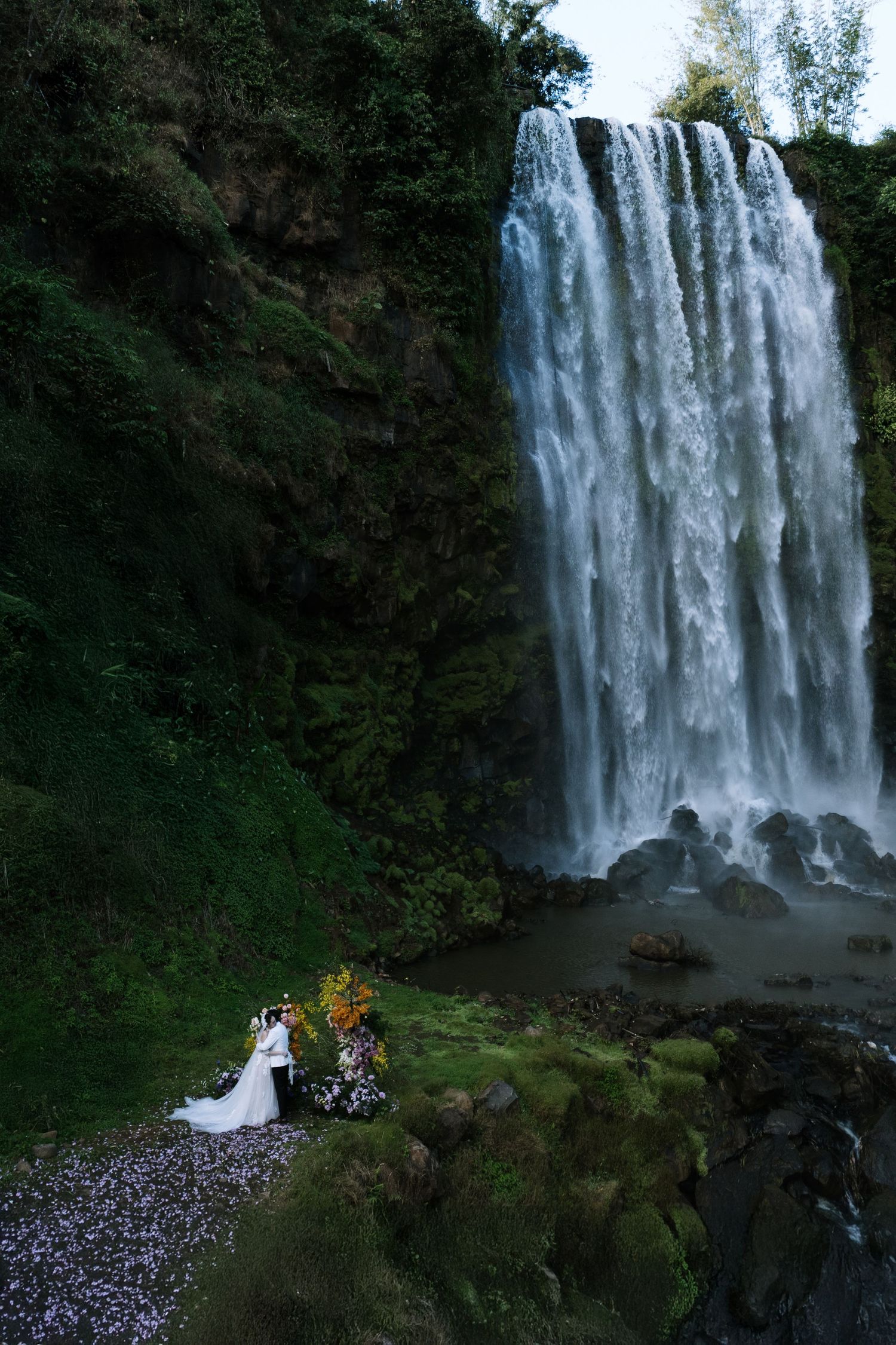 Bride and groom standing romantically before a massive cascading waterfall surrounded by green cliffs.
