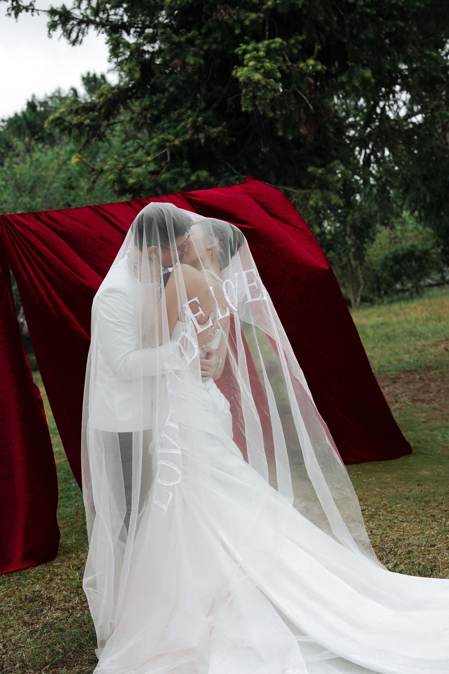 Bride and groom sharing a kiss hidden beneath a flowing veil near red fabric outdoors.