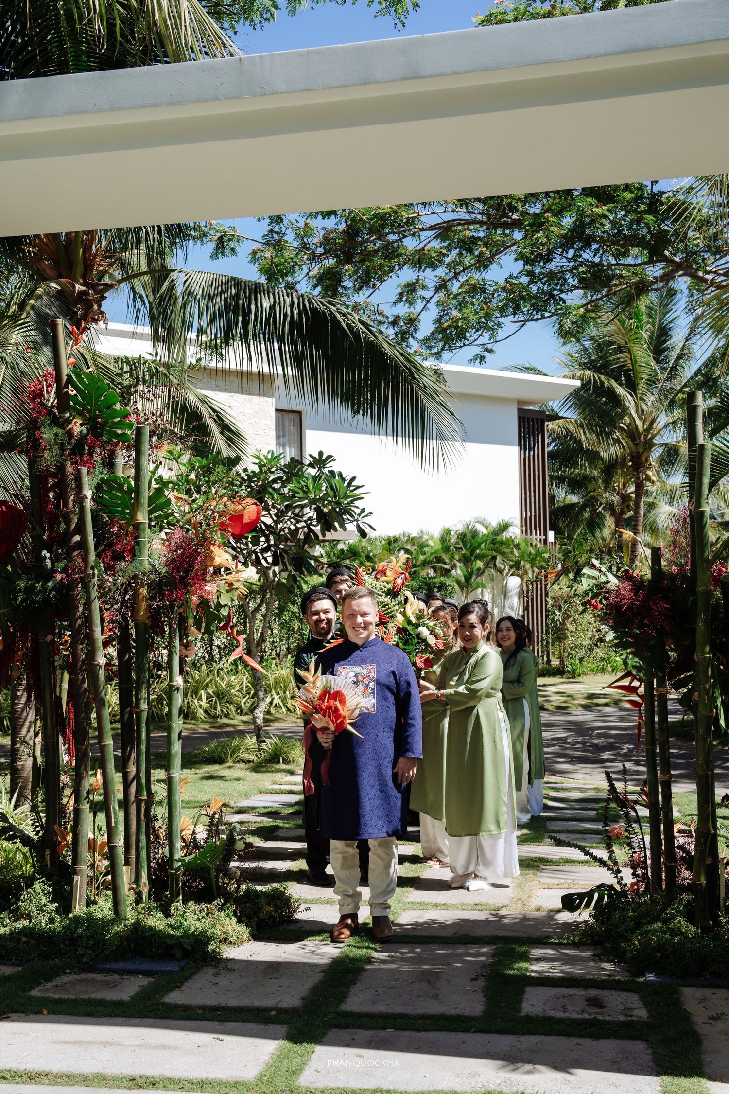 Group of people in traditional Vietnamese ao dai attire pose under a pergola surrounded by tropical garden.