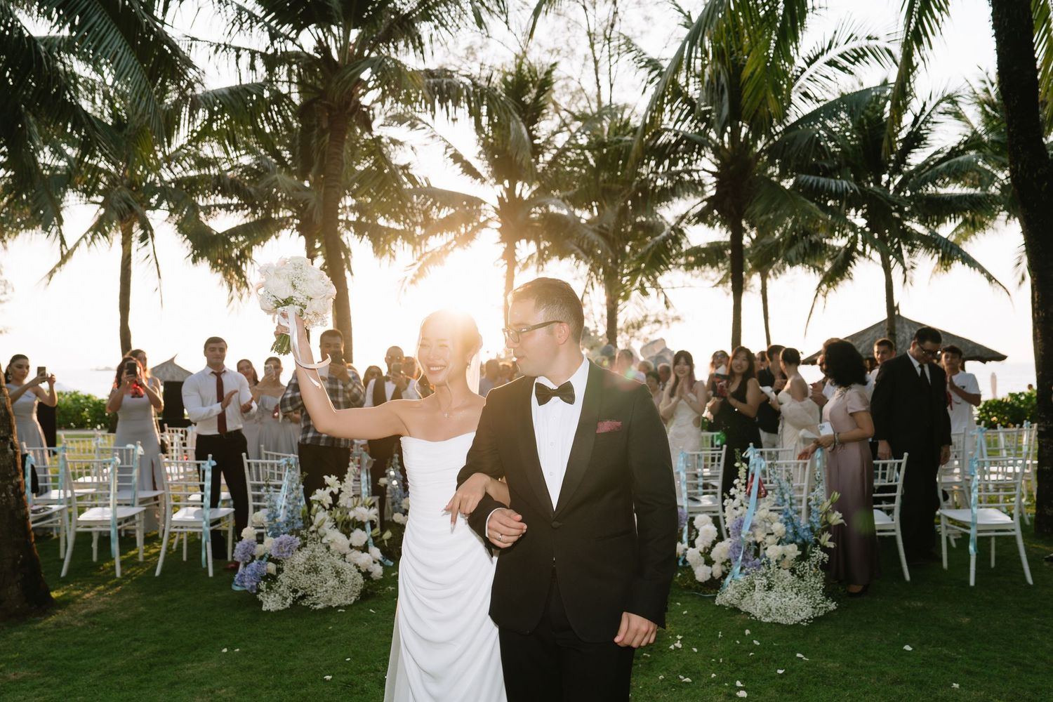 Joyful newlyweds exit outdoor ceremony under palm trees with golden sunset and cheering guests.