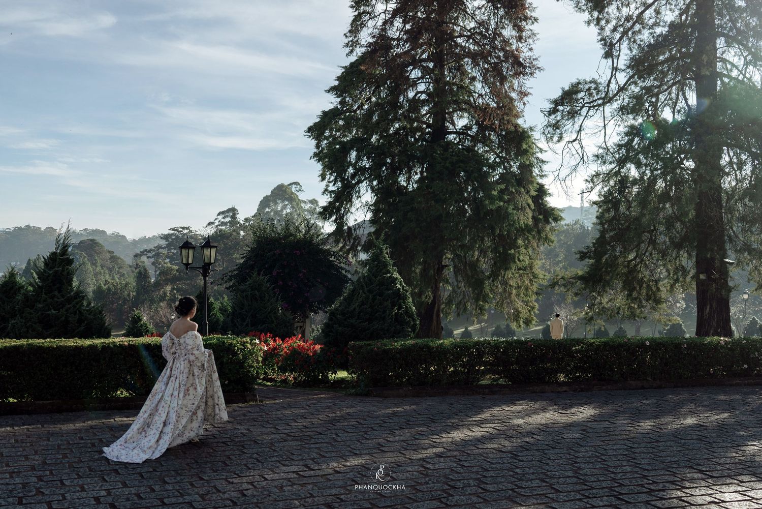 Bride in a floral gown twirls gracefully on a stone path surrounded by tall trees and manicured hedges.