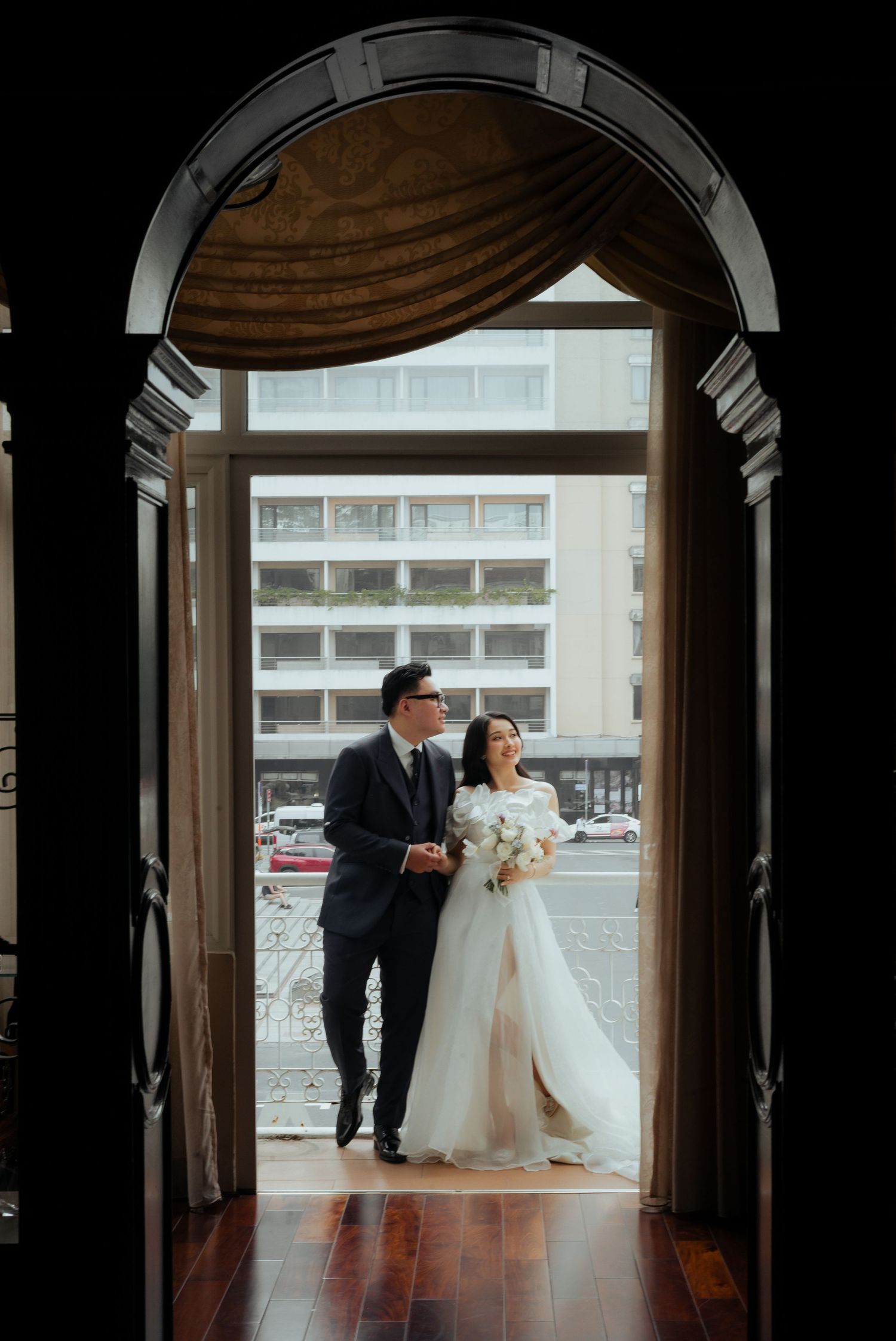 Elegant couple in wedding attire stand framed by a grand arched doorway with city views behind them.