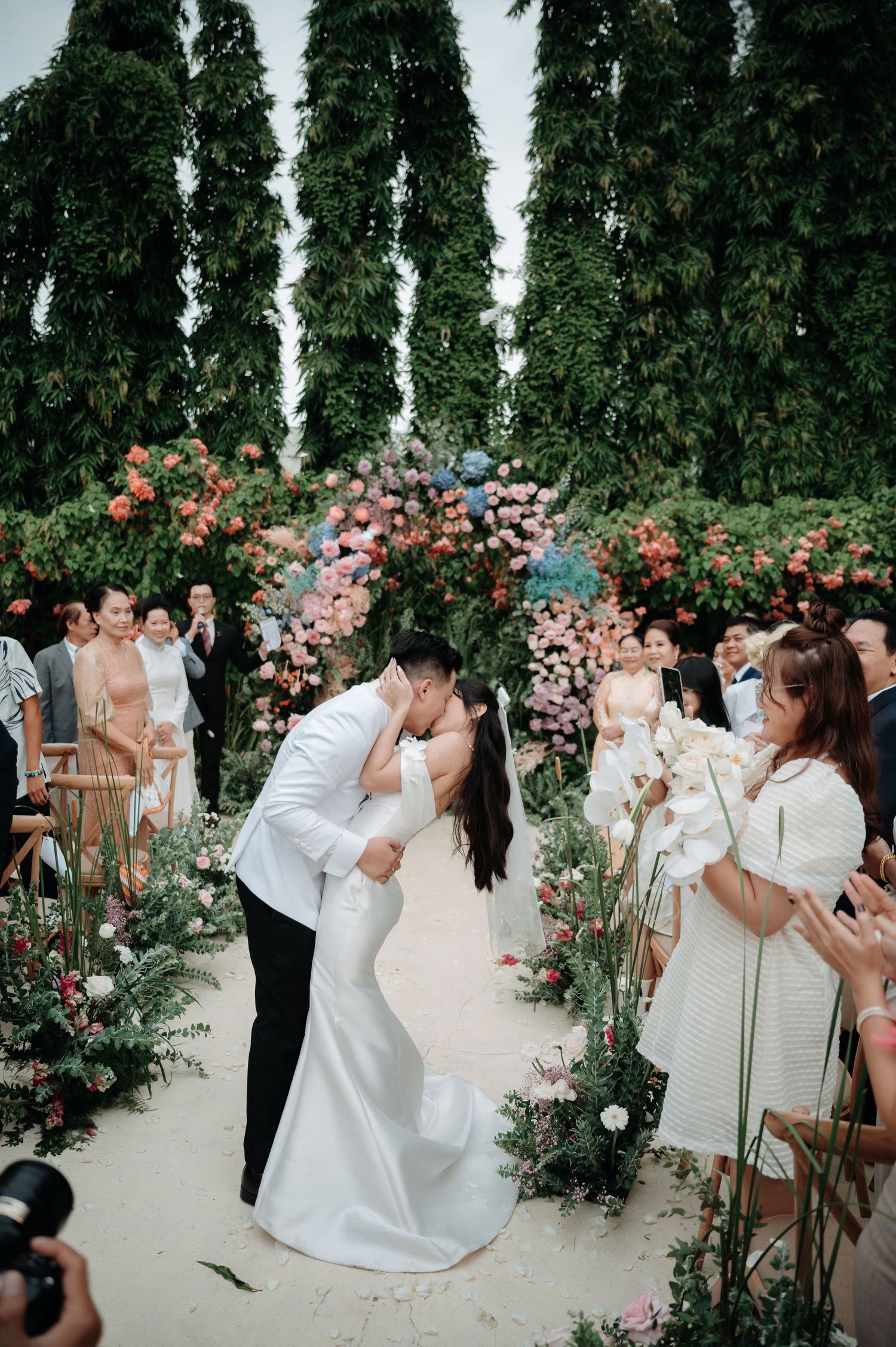 Bride and groom share a romantic kiss at outdoor ceremony beneath a lush floral arch with guests watching.