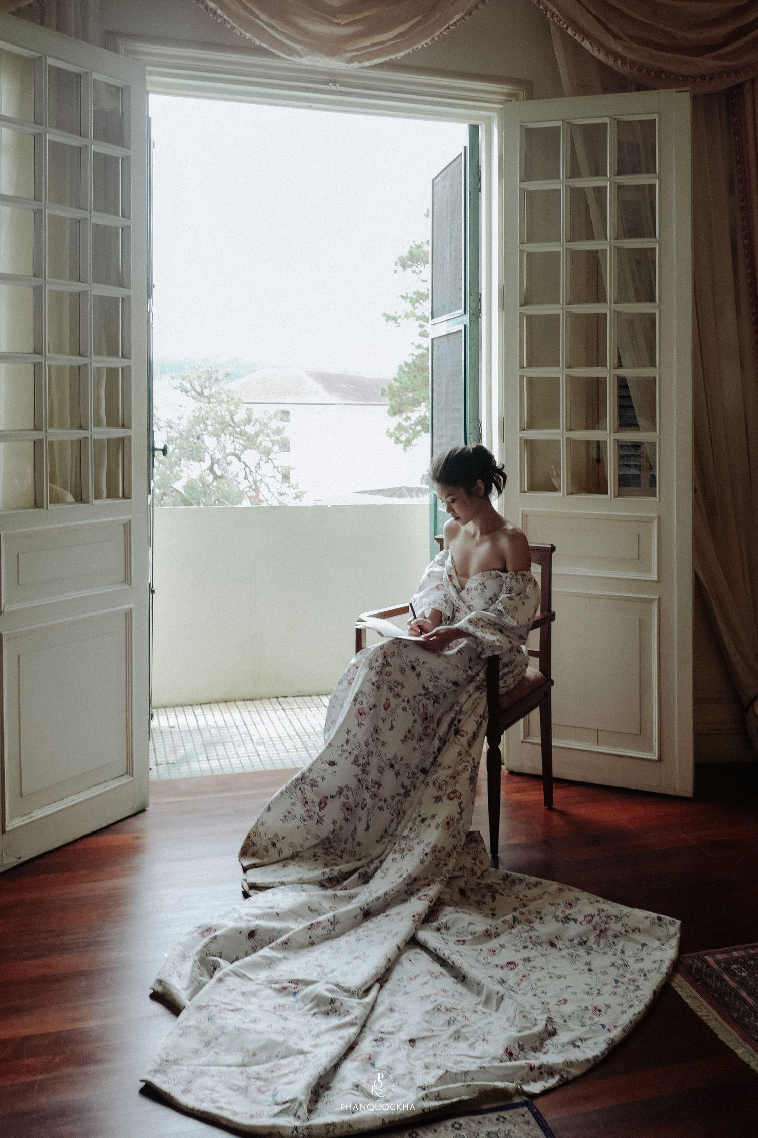 Bride in a floral off-shoulder gown sits by open French doors reading in soft natural light.