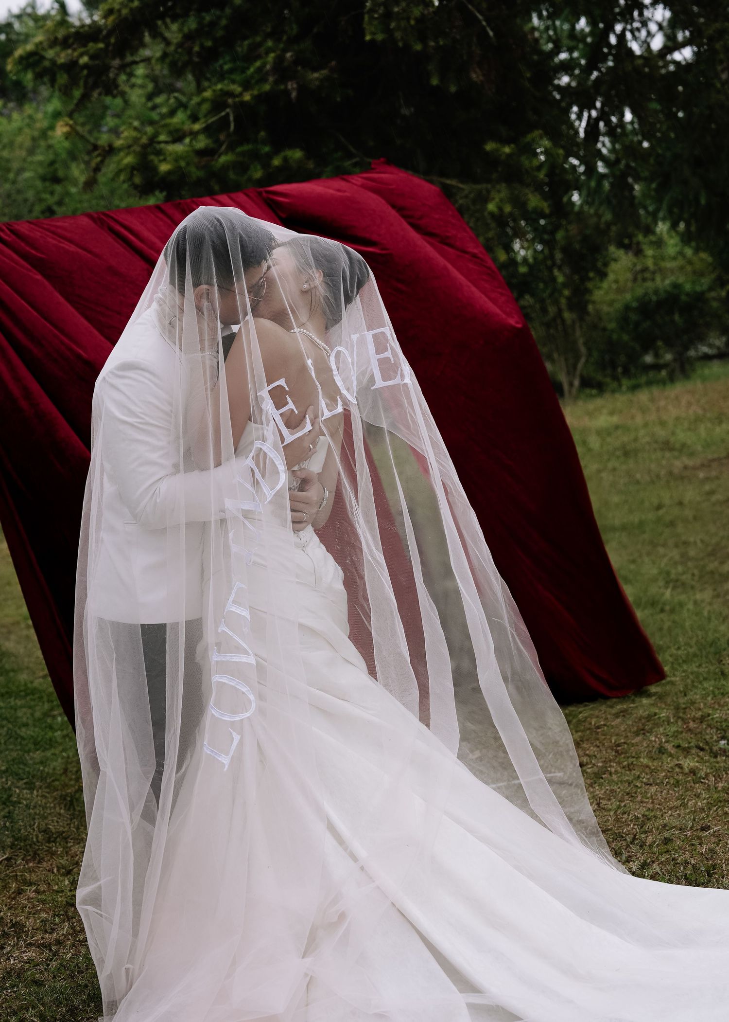 Bride and groom sharing a kiss under an embroidered wedding veil in front of a red velvet backdrop outdoors.