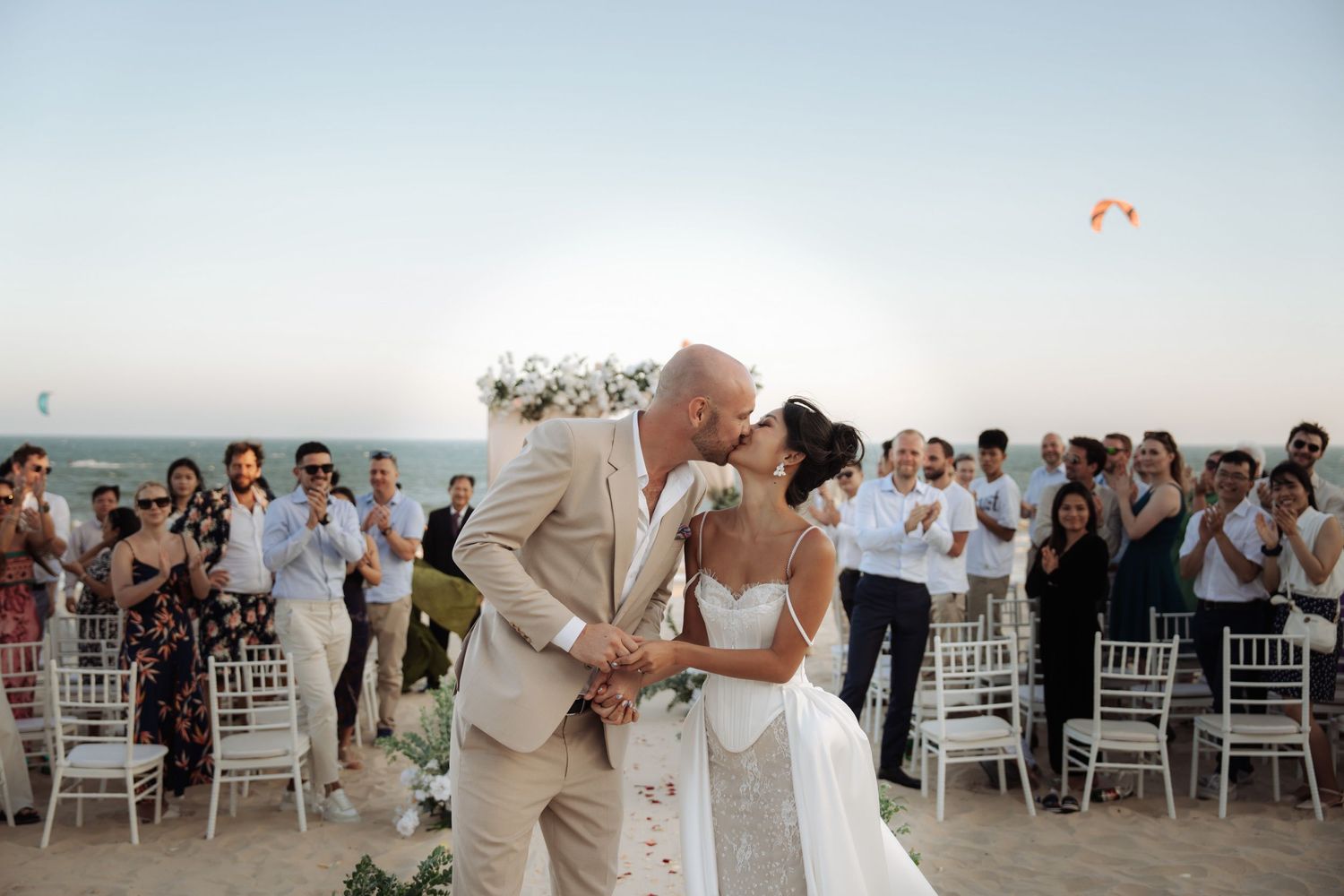 Bride and groom share first kiss at outdoor beach wedding ceremony surrounded by applauding guests at sunset.