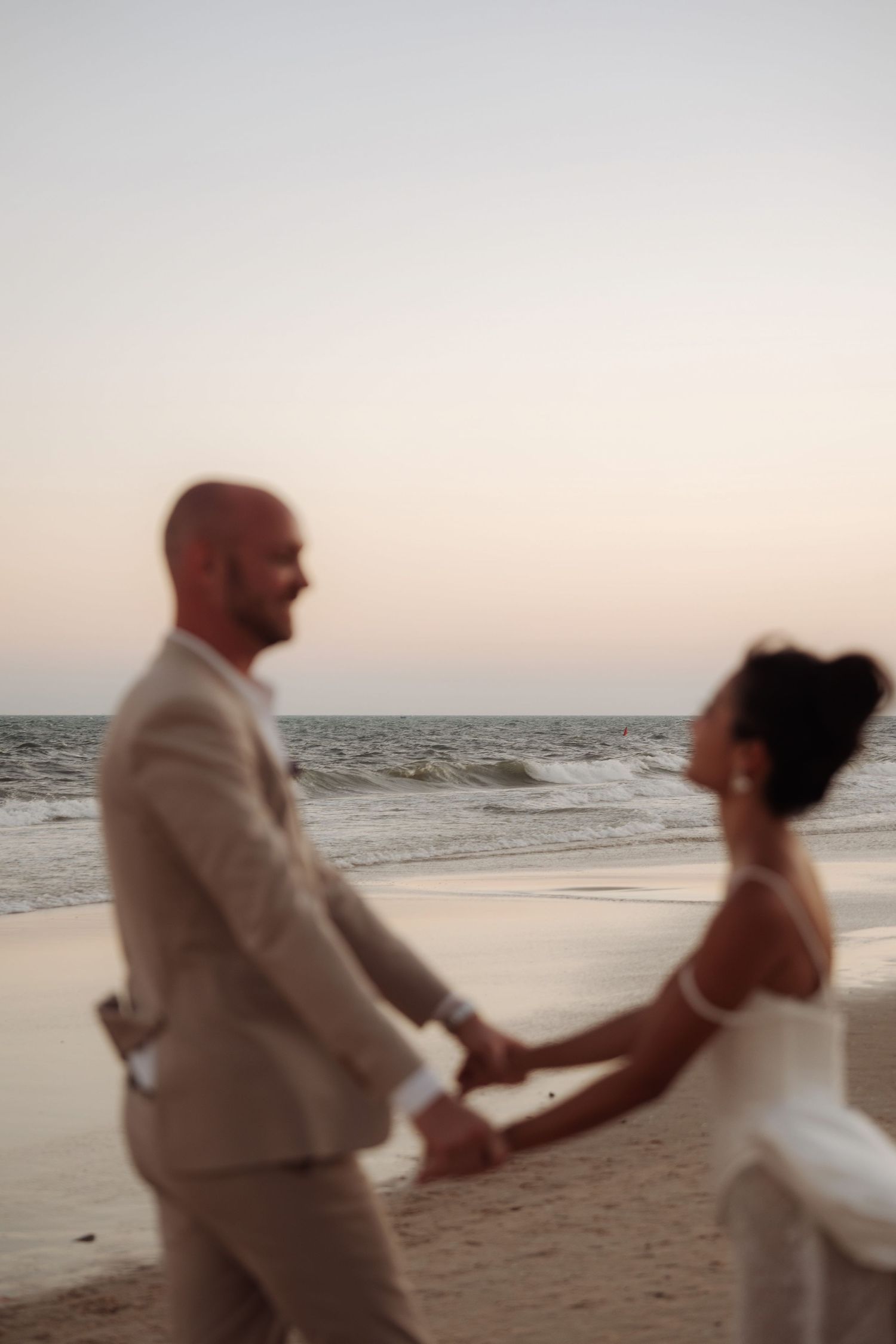 Bride and groom hold hands walking along a serene beach at sunset during their wedding day.