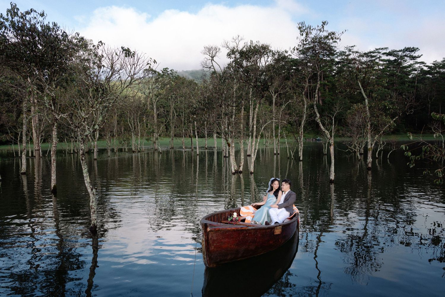 Romantic couple sitting together in a wooden rowboat on a calm lake surrounded by lush trees.