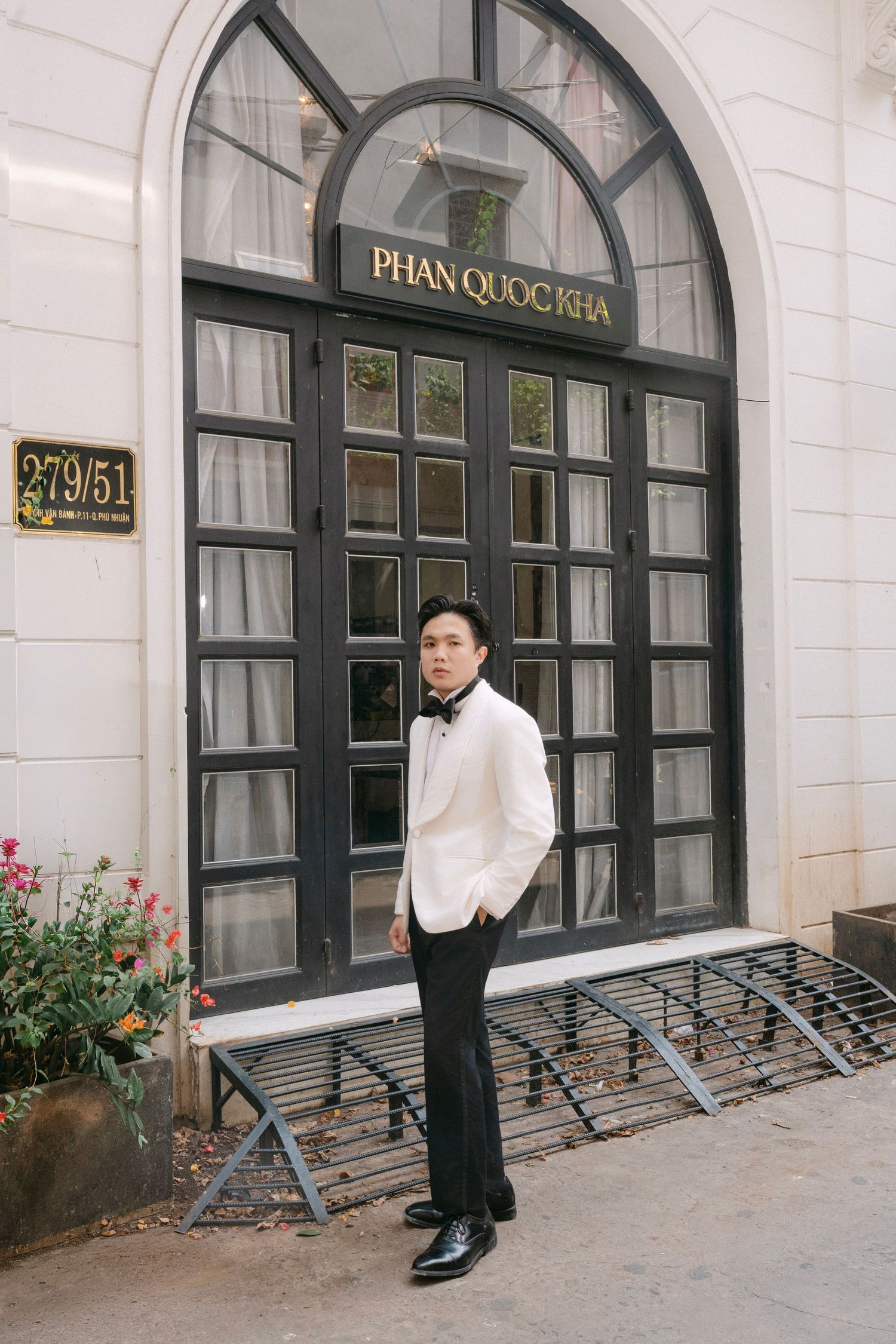Groom in white tuxedo standing before Phan Quoc Kha boutique entrance with arched glass doors.