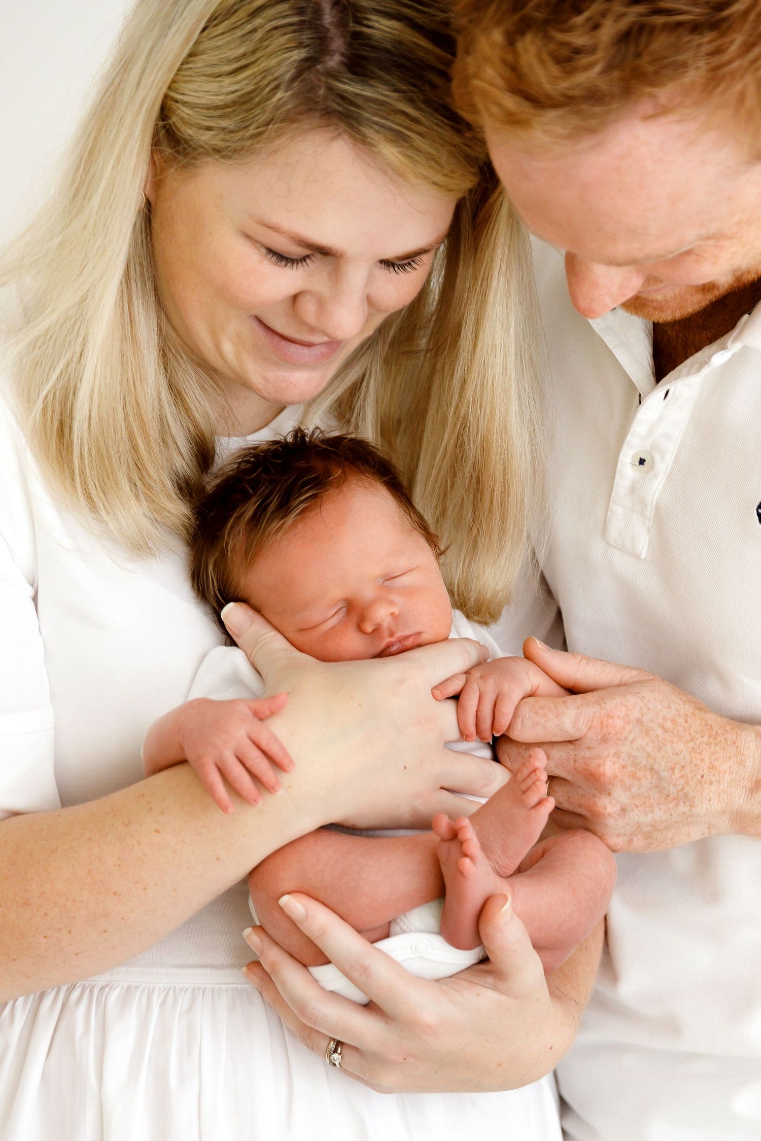 Parents in white clothing gently hold and admire their sleeping newborn baby.