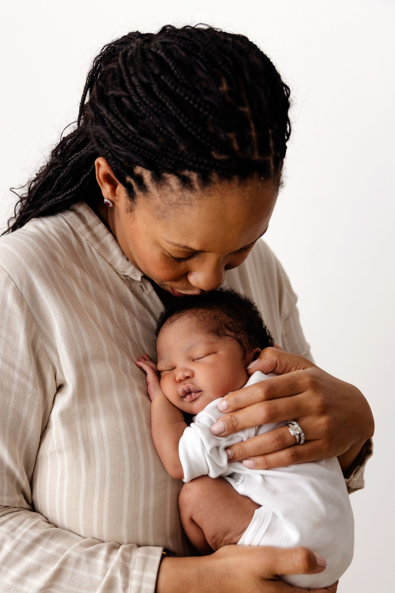 A tender moment between a mother in beige sweater cradling her sleeping newborn baby against her chest.