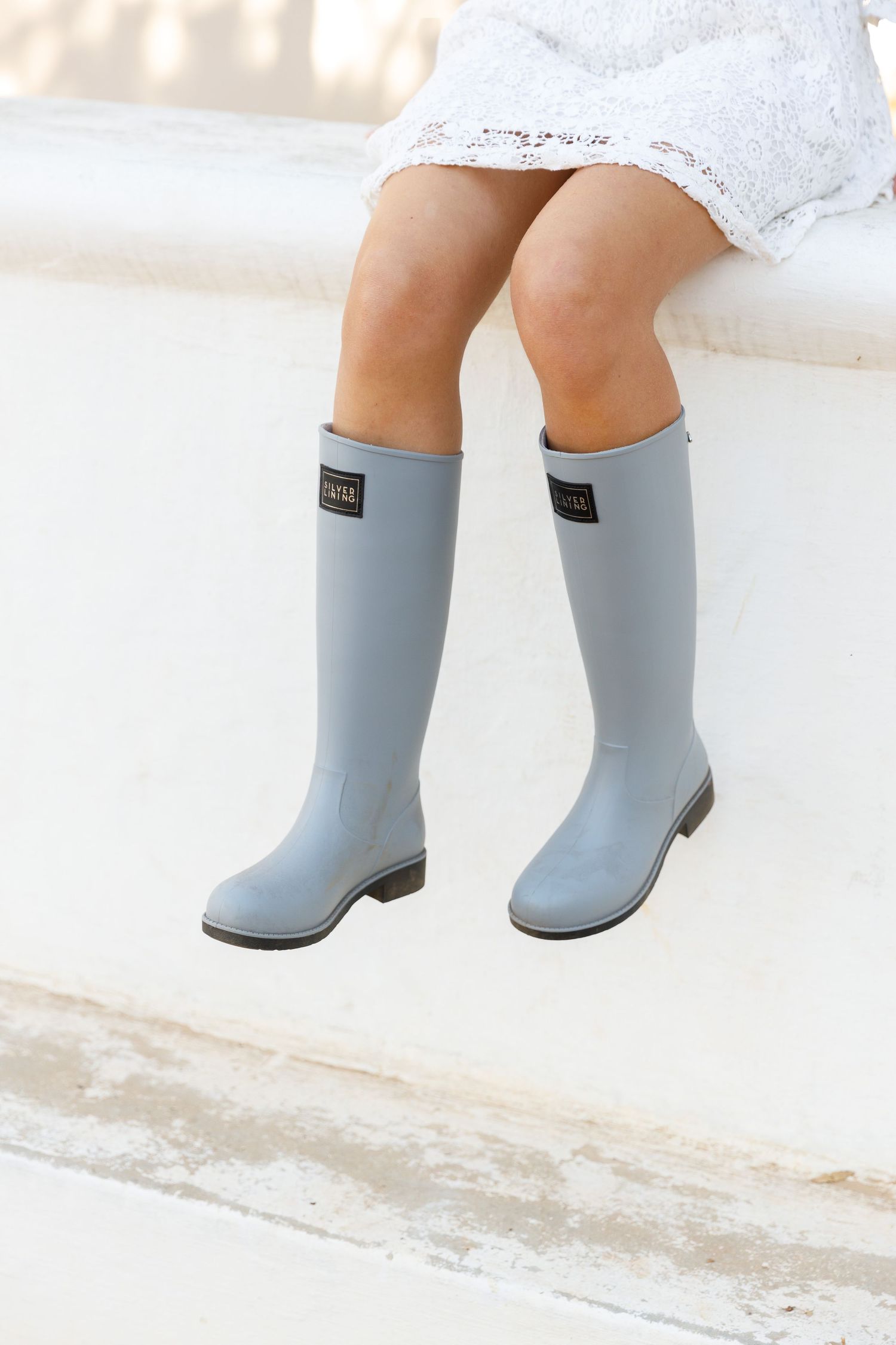 A person wearing light gray rubber rain boots and a white dress sits on a light colored surface, legs dangling.