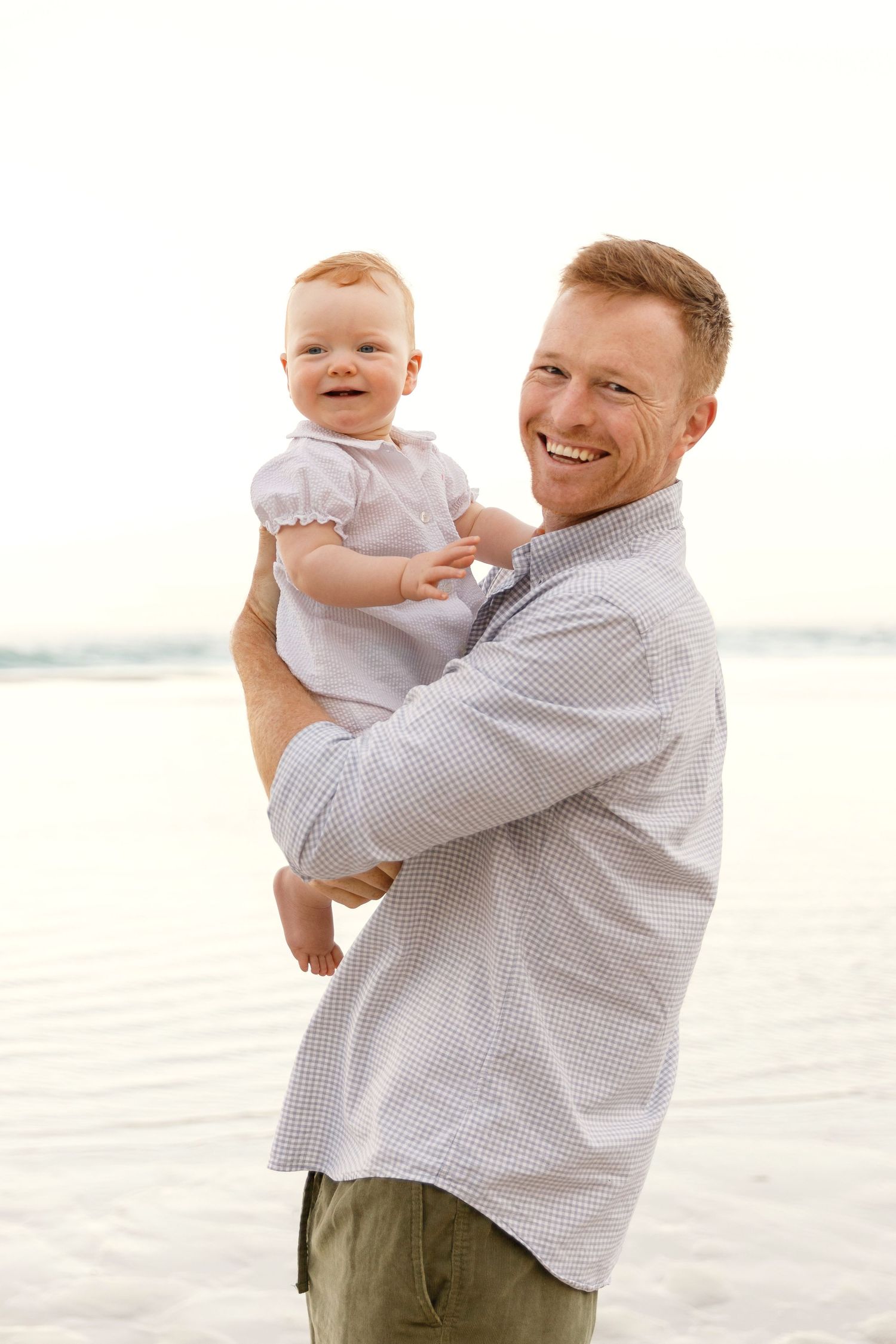A baby in a white dress smiles while being held by someone in a light striped shirt on a bright sunny day.