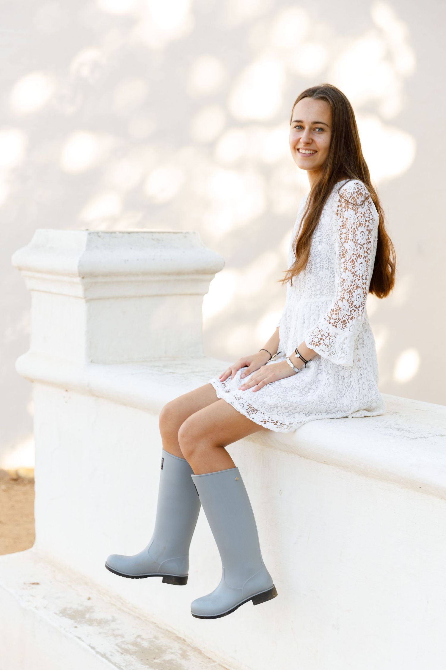 Person in white lace dress and gray rain boots sits on white stone bench in soft sunlit setting.