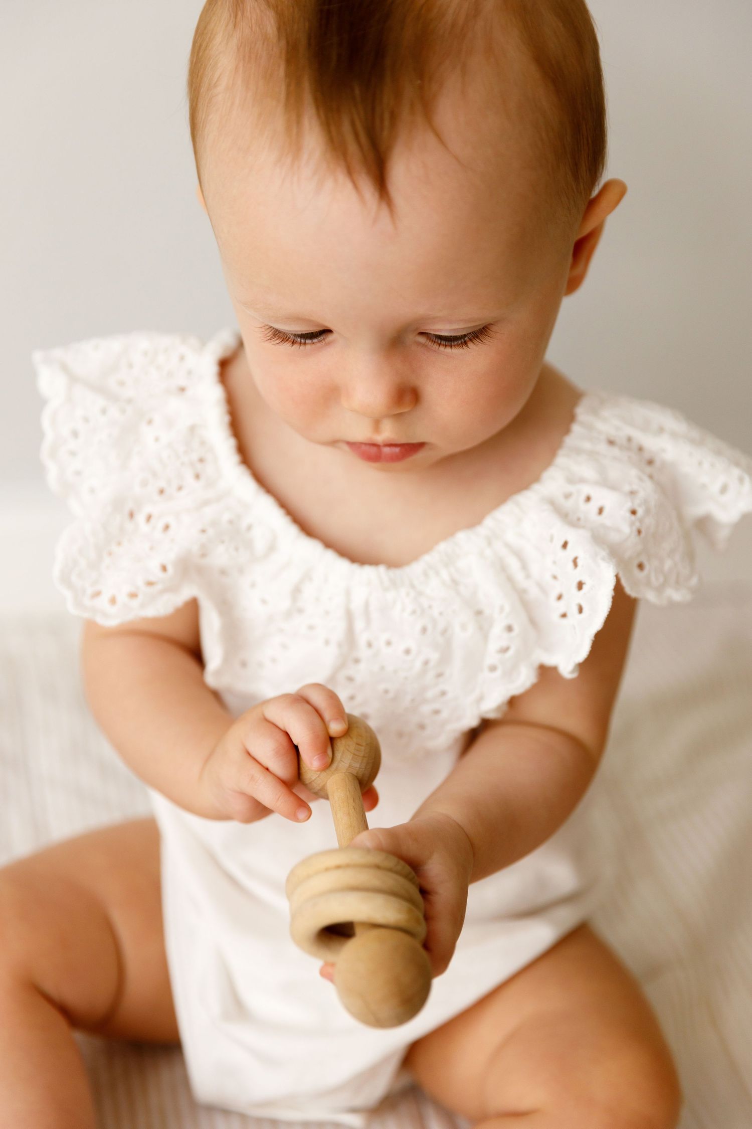 Baby in white eyelet lace dress holds wooden rattle toy while looking down against neutral background.