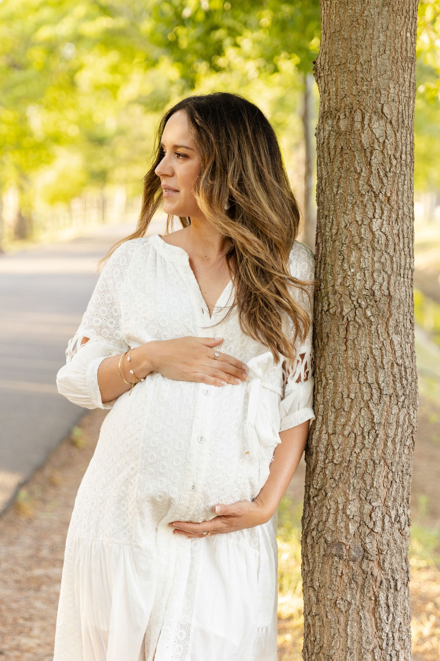 A pregnant woman in a flowing white dress stands next to a tree trunk in dappled sunlight on an autumn day.