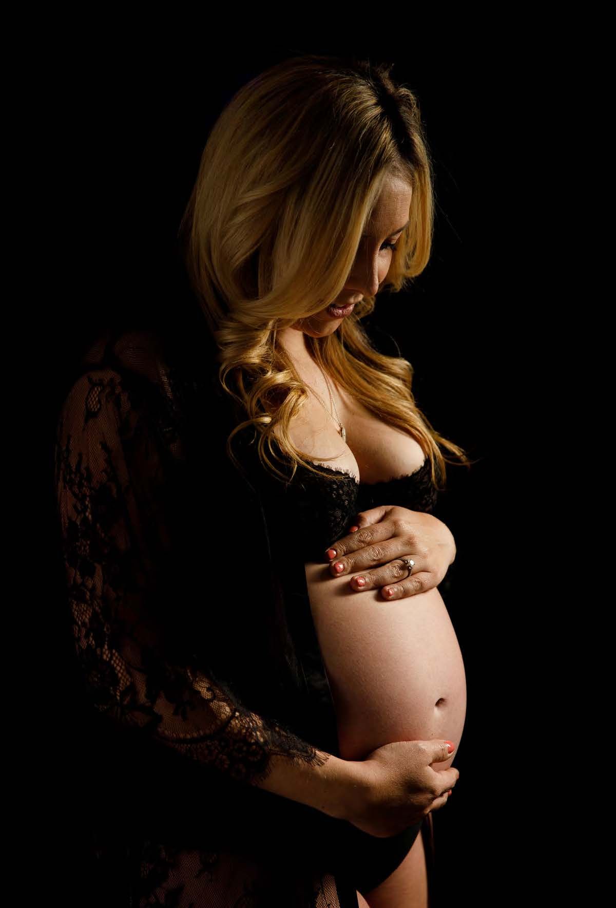 Silhouetted figure in black lace gently caresses pregnant belly in artistic maternity photo against dark background.