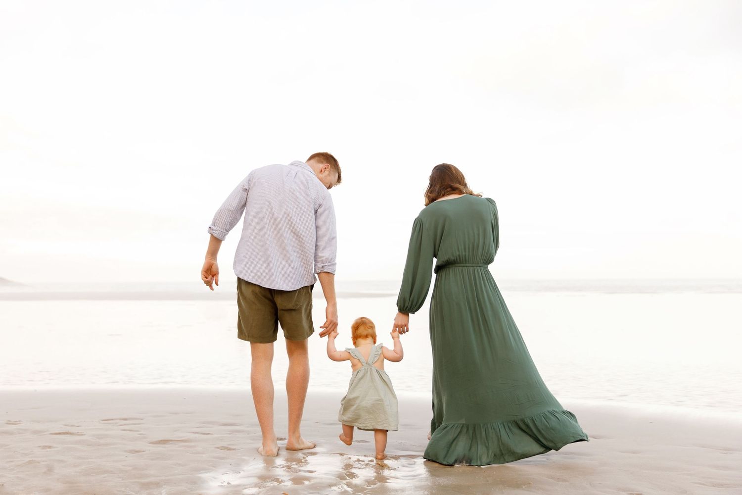 Family of three holding hands while walking together on a sandy beach at sunset in soft neutral colors.