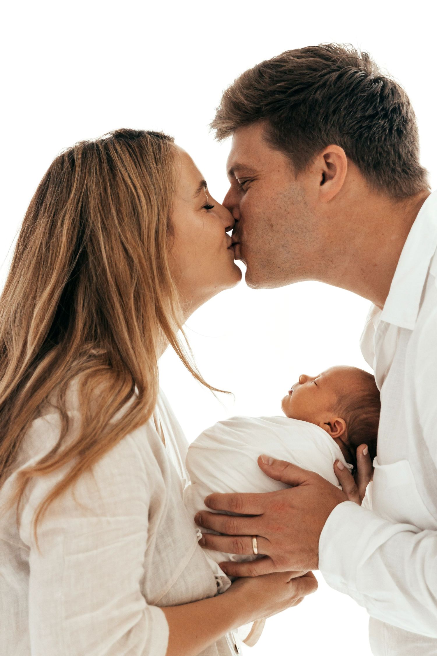 A couple shares a tender kiss while holding their newborn baby against a white background.