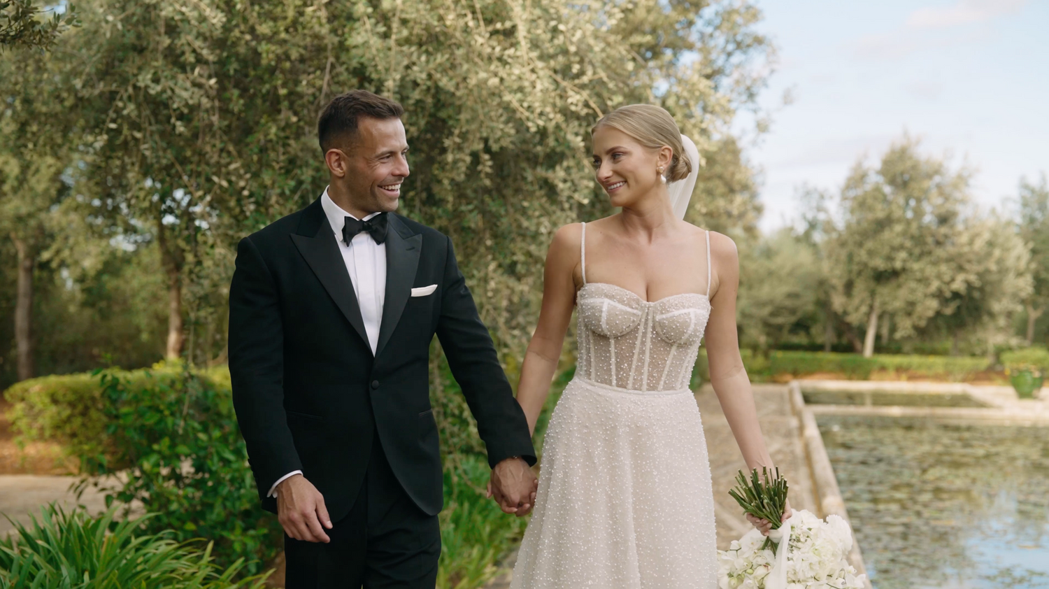 Bride and groom walking hand in hand through a garden with a pond, looking at each other with joy and complicity.