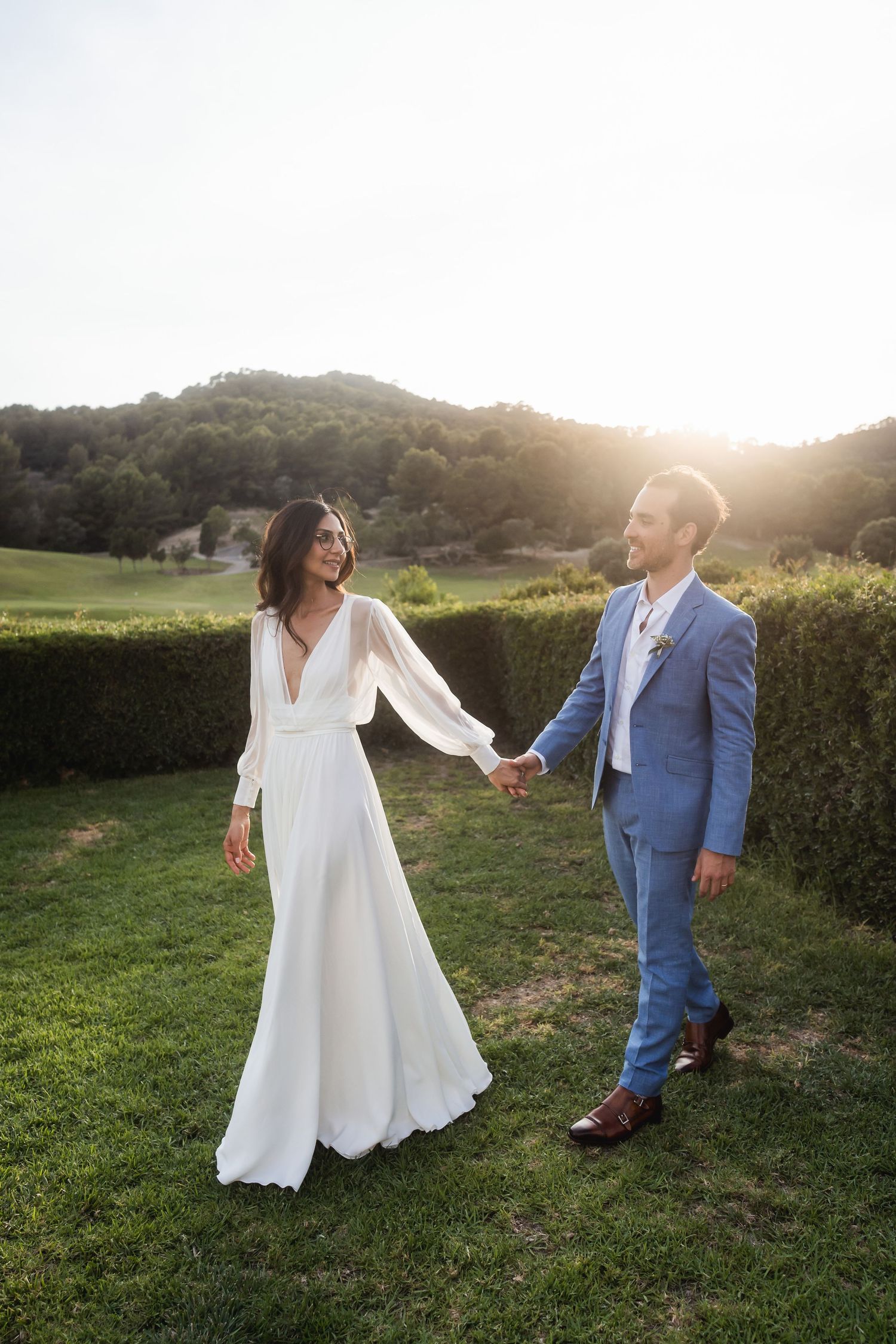 Newlywed couple walking hand in hand through a garden at sunset, enjoying a special moment.