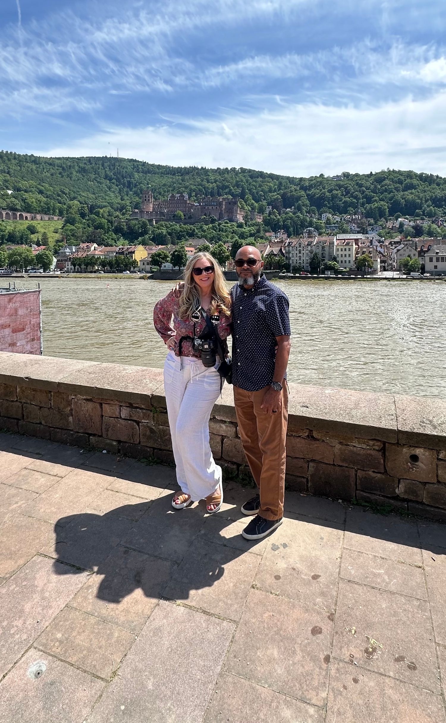 Tourists pose along the river in Heidelberg with green hills and historic architecture visible in the background.