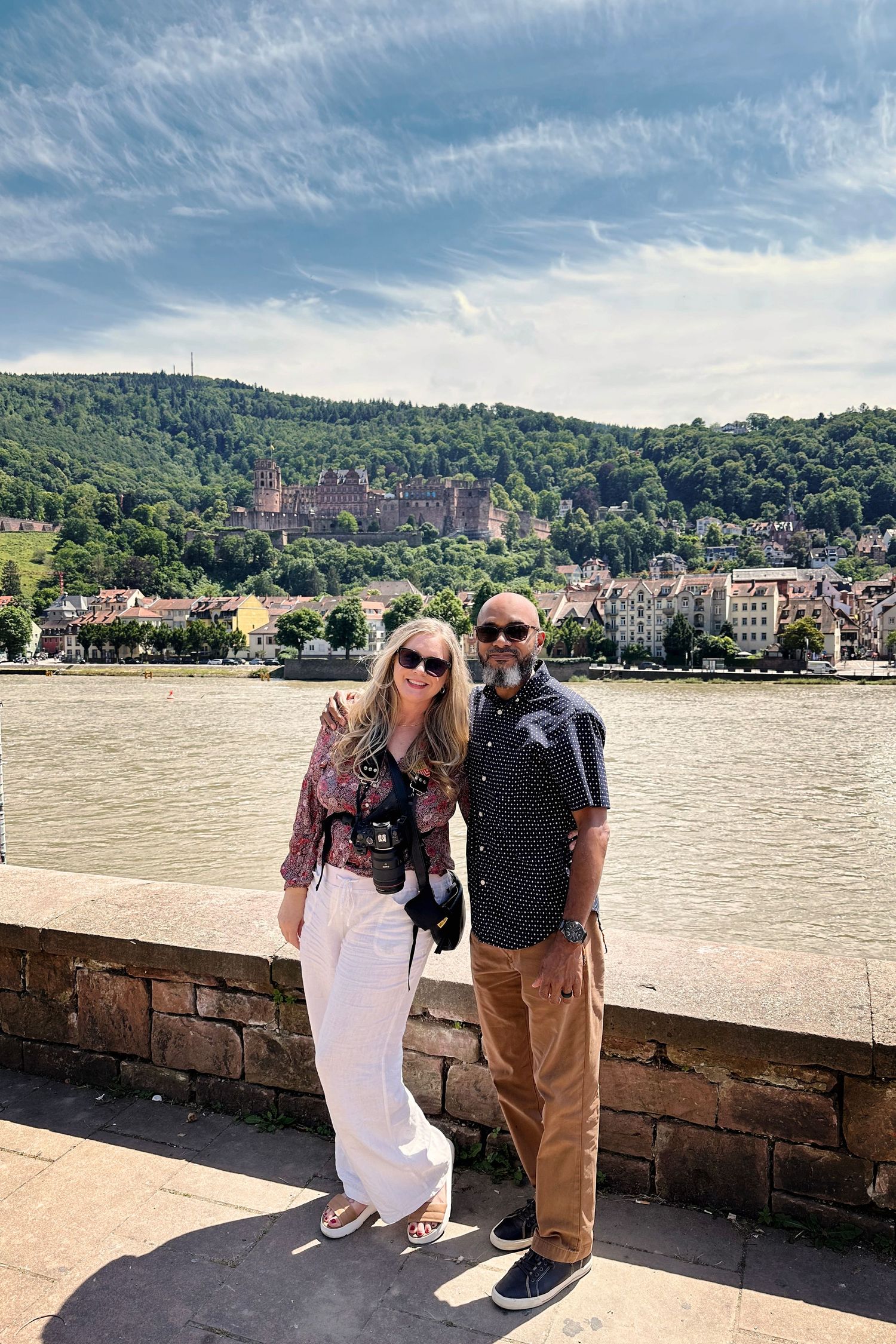 Tourists posing along stone wall by river with historic castle and hillside buildings of Heidelberg in background.