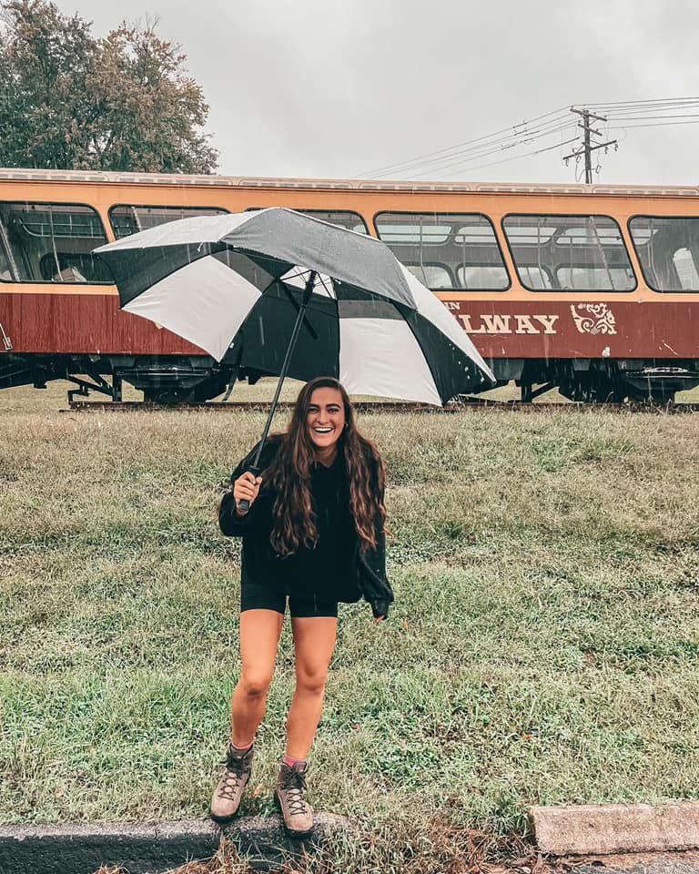 Person with umbrella poses in front of vintage orange train cars on rainy day.