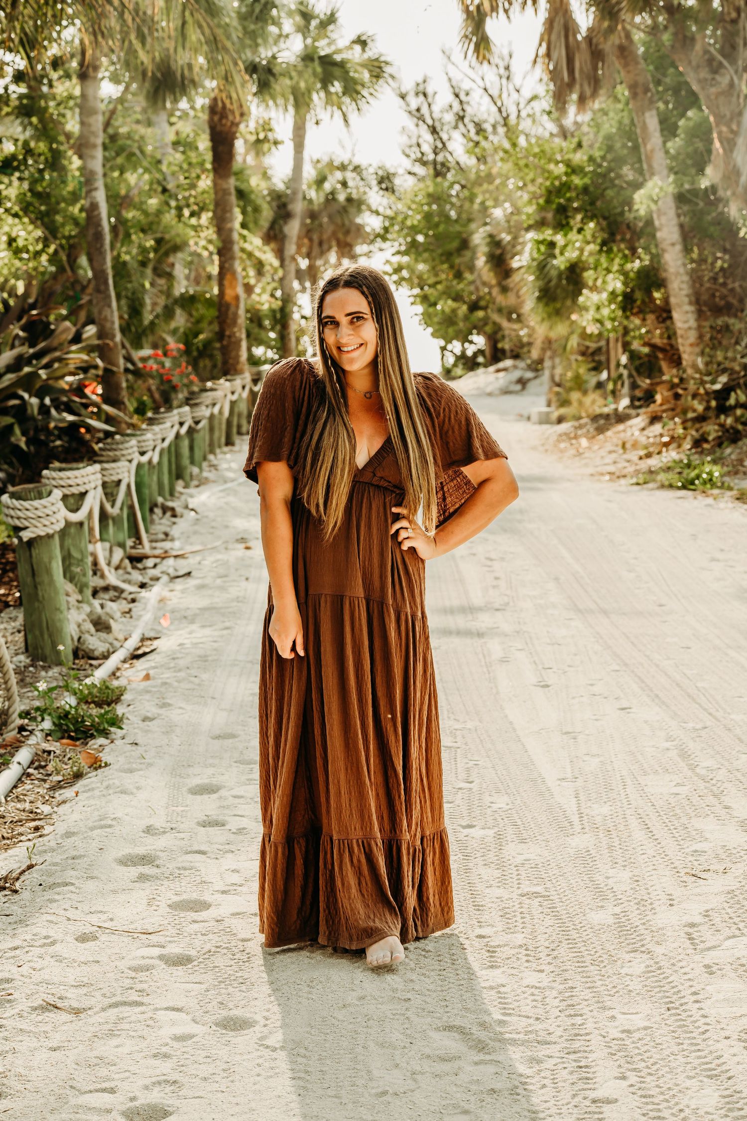 Woman in flowing brown maxi dress poses on sandy palm-lined pathway.