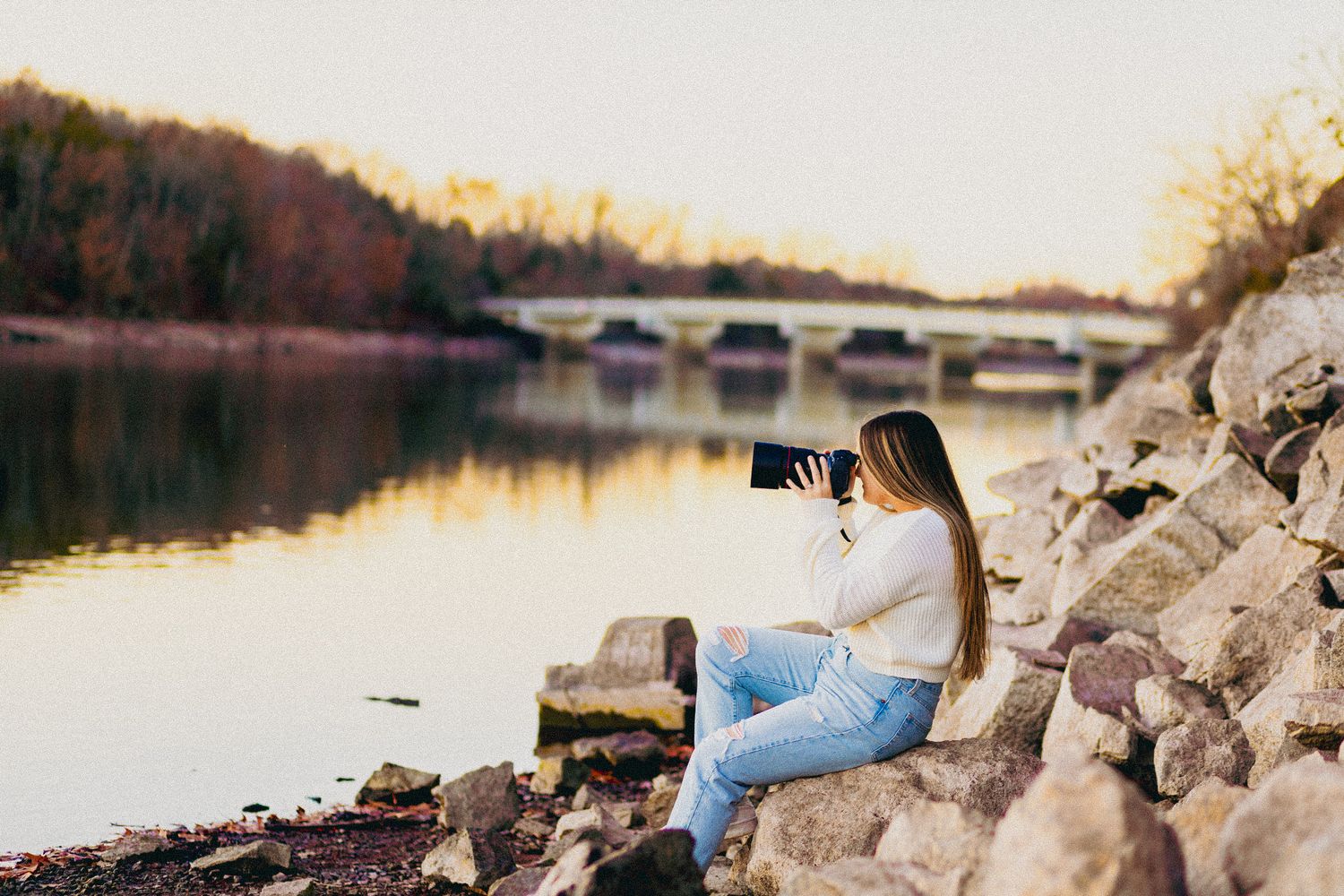 Photographer sitting on rocky shoreline capturing sunset over lake and bridge.