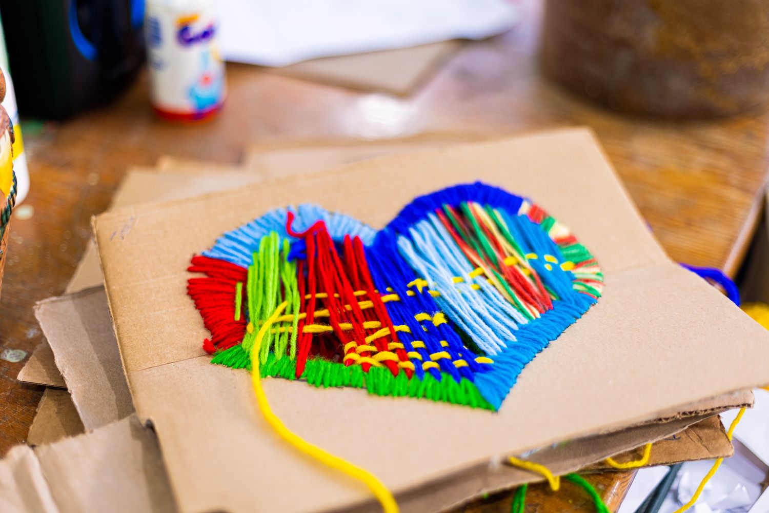 Colorful heart made from interwoven yarn strips on brown cardboard background.