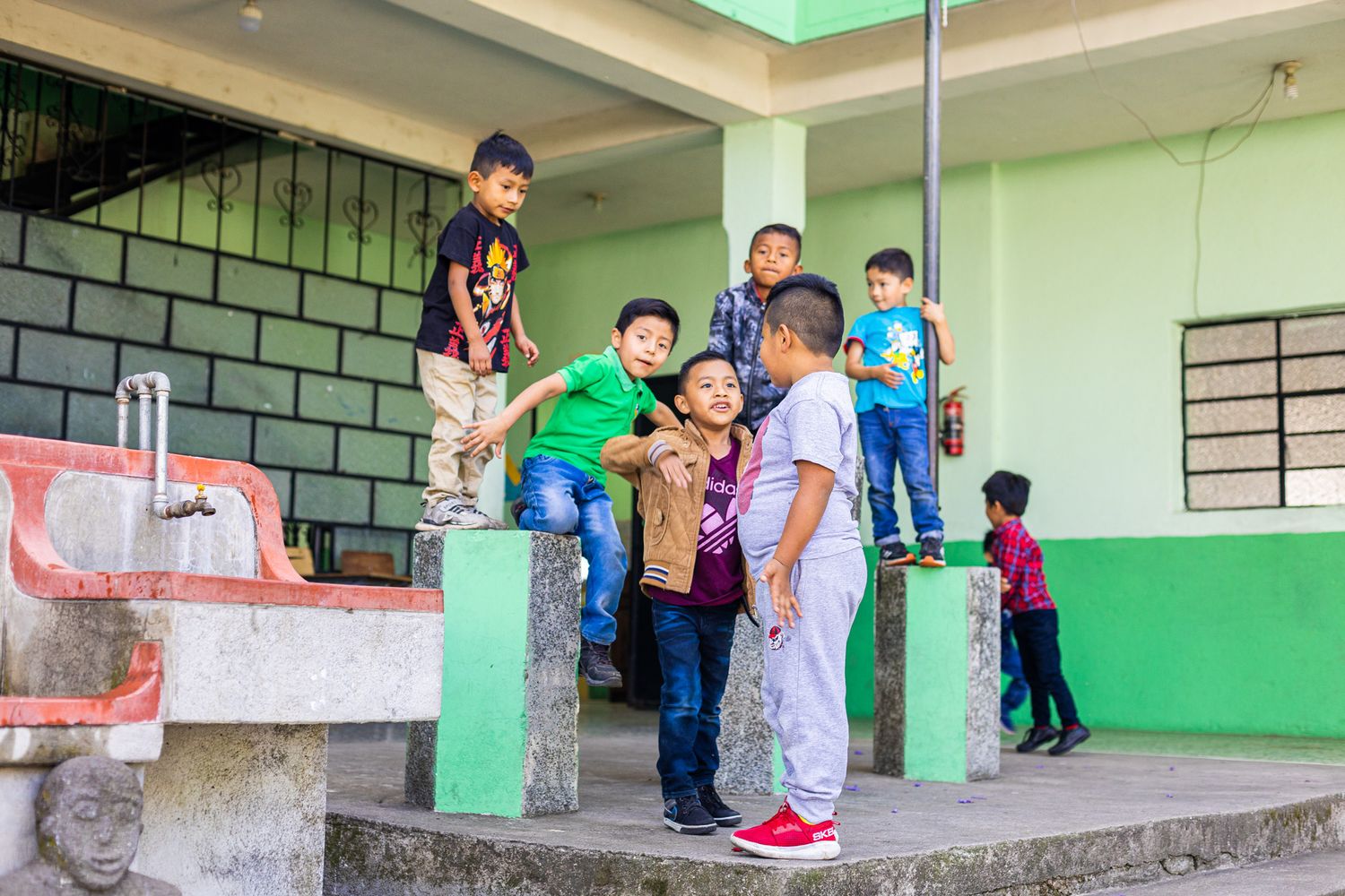 Children and adults socialize on the steps of a green-painted school building.