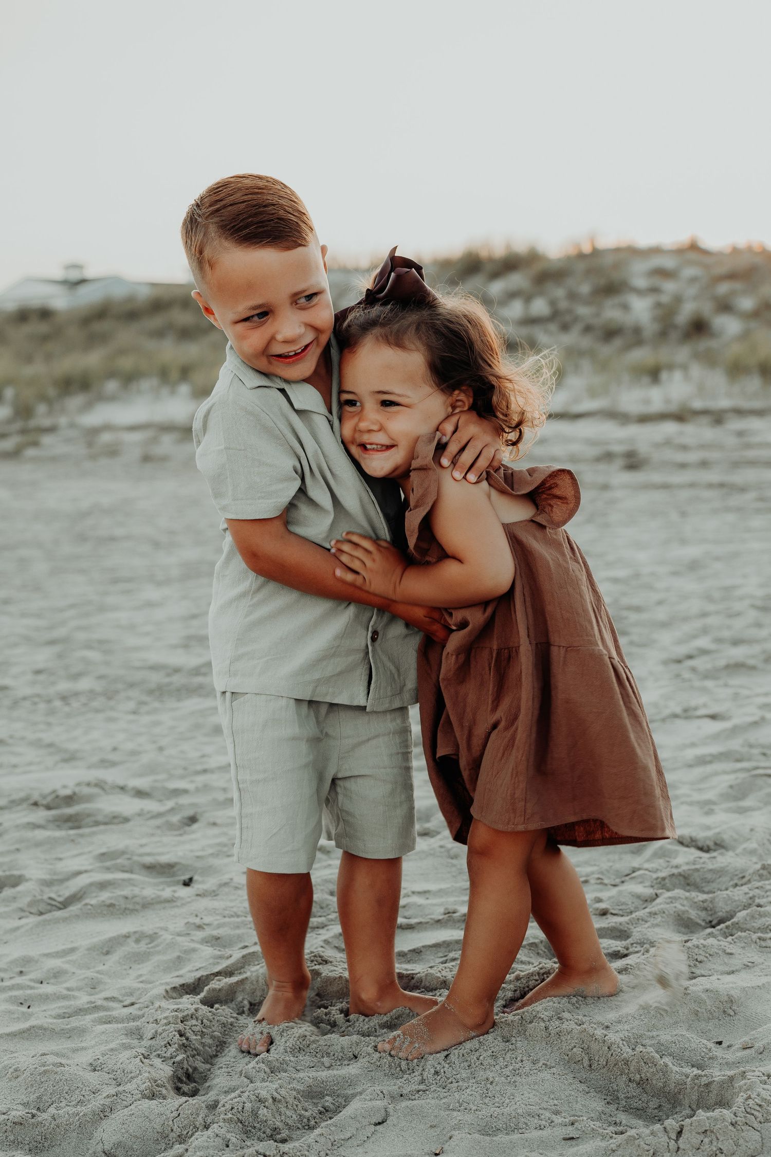 Two young children in beige and brown outfits play and laugh together on a sandy beach.