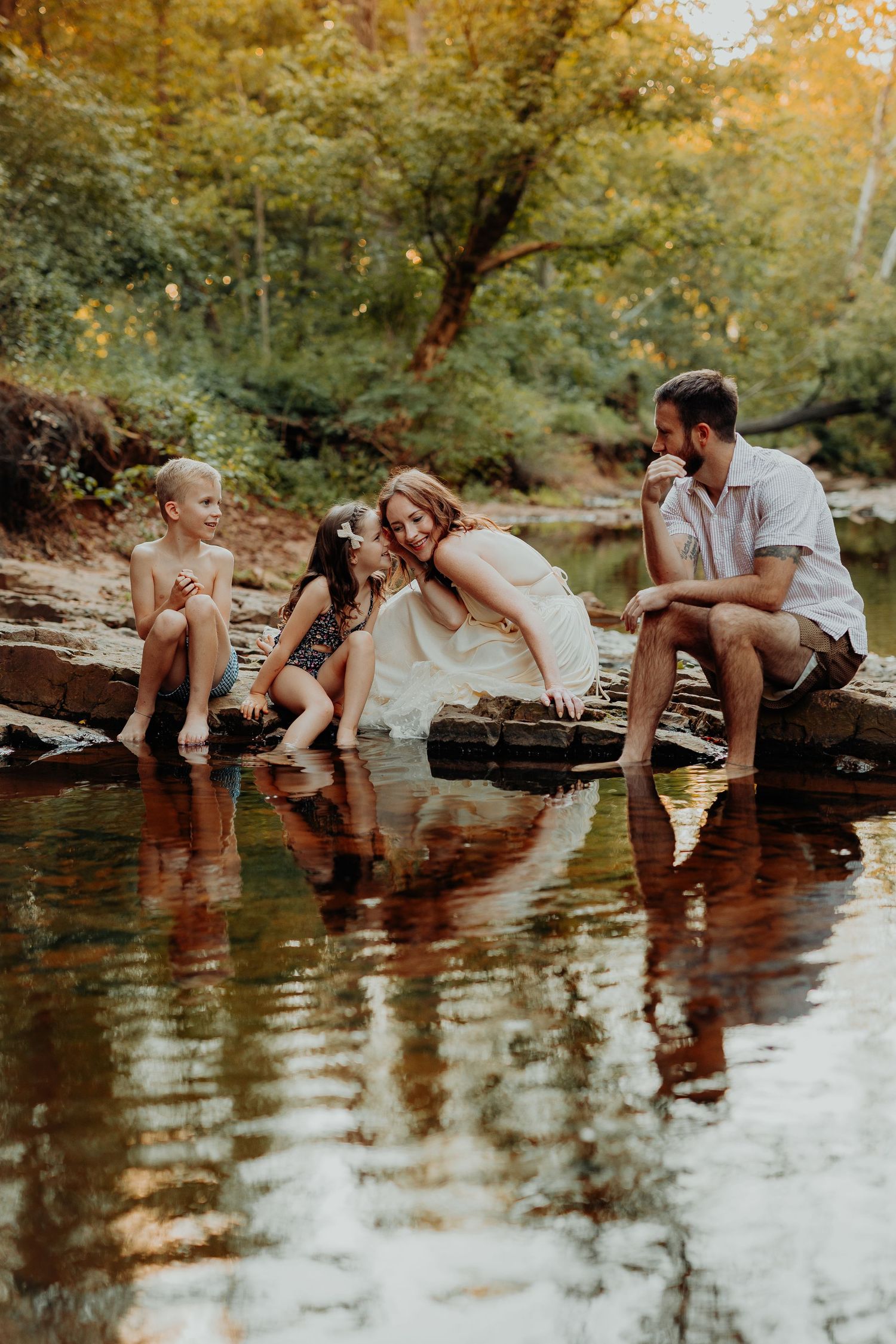 A romantic couple sits on a fallen log over a creek with their reflections visible in the calm water at sunset.