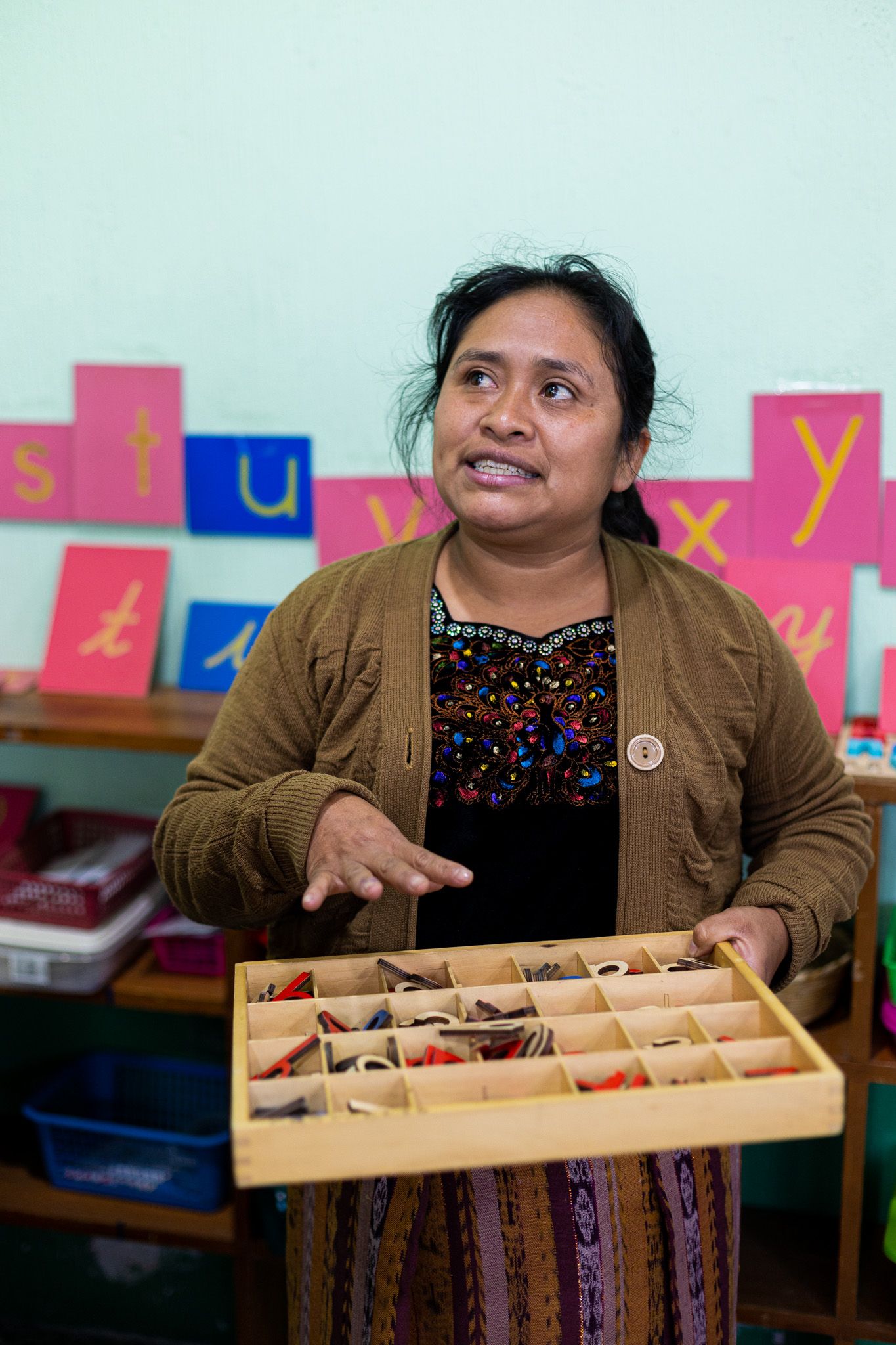 Woman in traditional dress holding a display box in classroom with alphabet letters on wall.