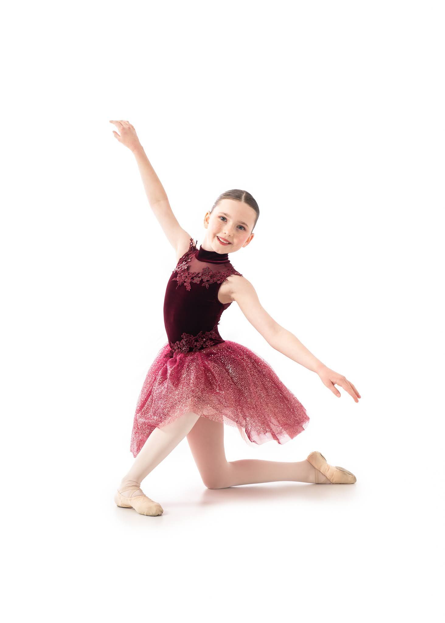 Young ballet dancer poses gracefully in burgundy velvet and lace tutu costume on white background.