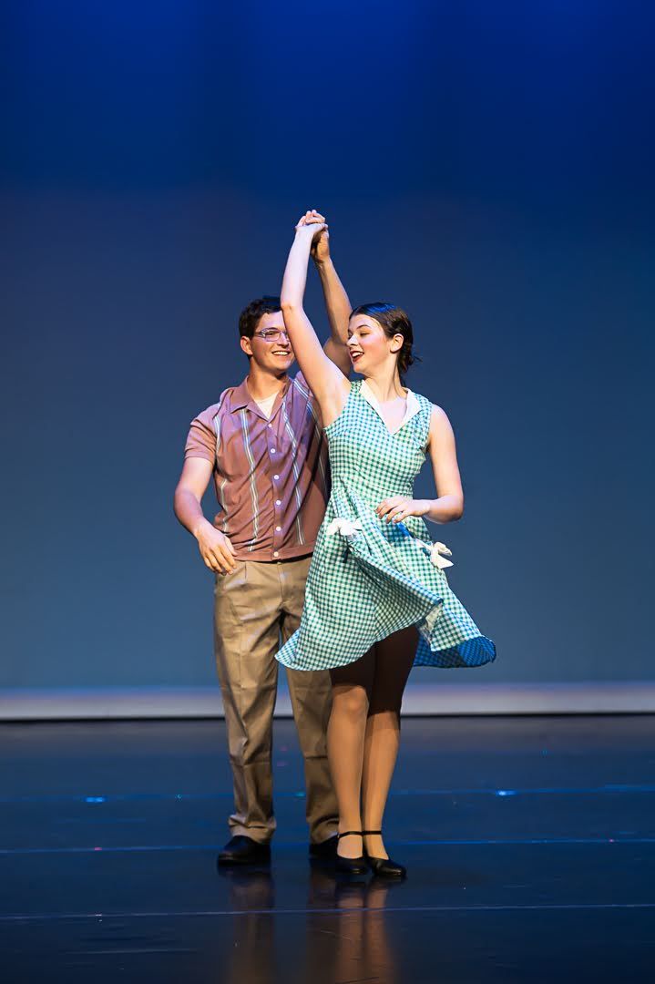 A smiling couple dances together on stage, the woman in a green gingham dress being twirled by her partner.