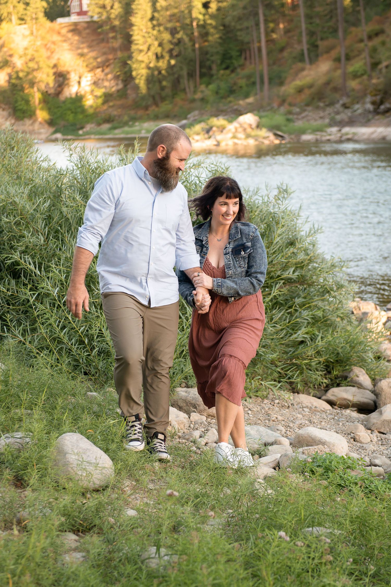 Happy couple holding hands walking along rocky riverbank with lush greenery and water in background.