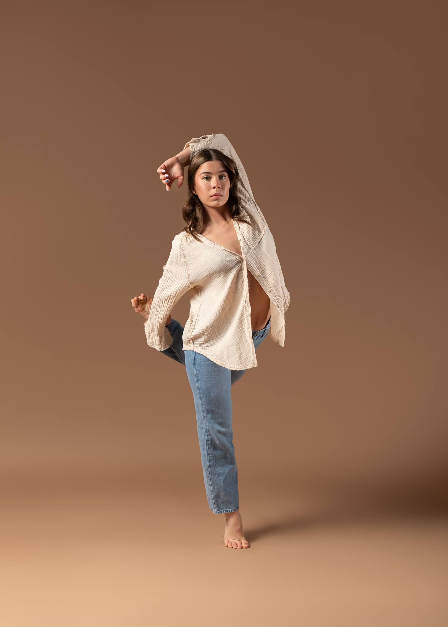 Young dancer in casual jeans and cream top performs hip-hop move on warm brown background.